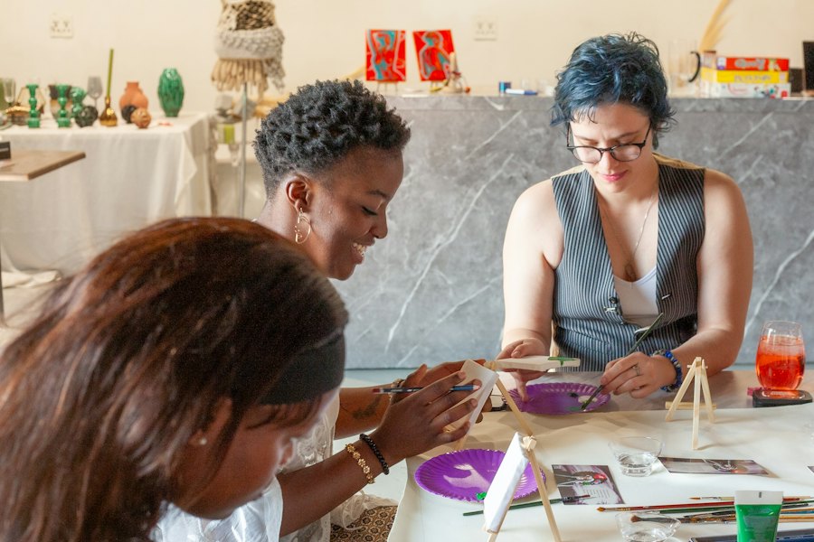 Três mulheres pintam e montam peças numa mesa de oficina.