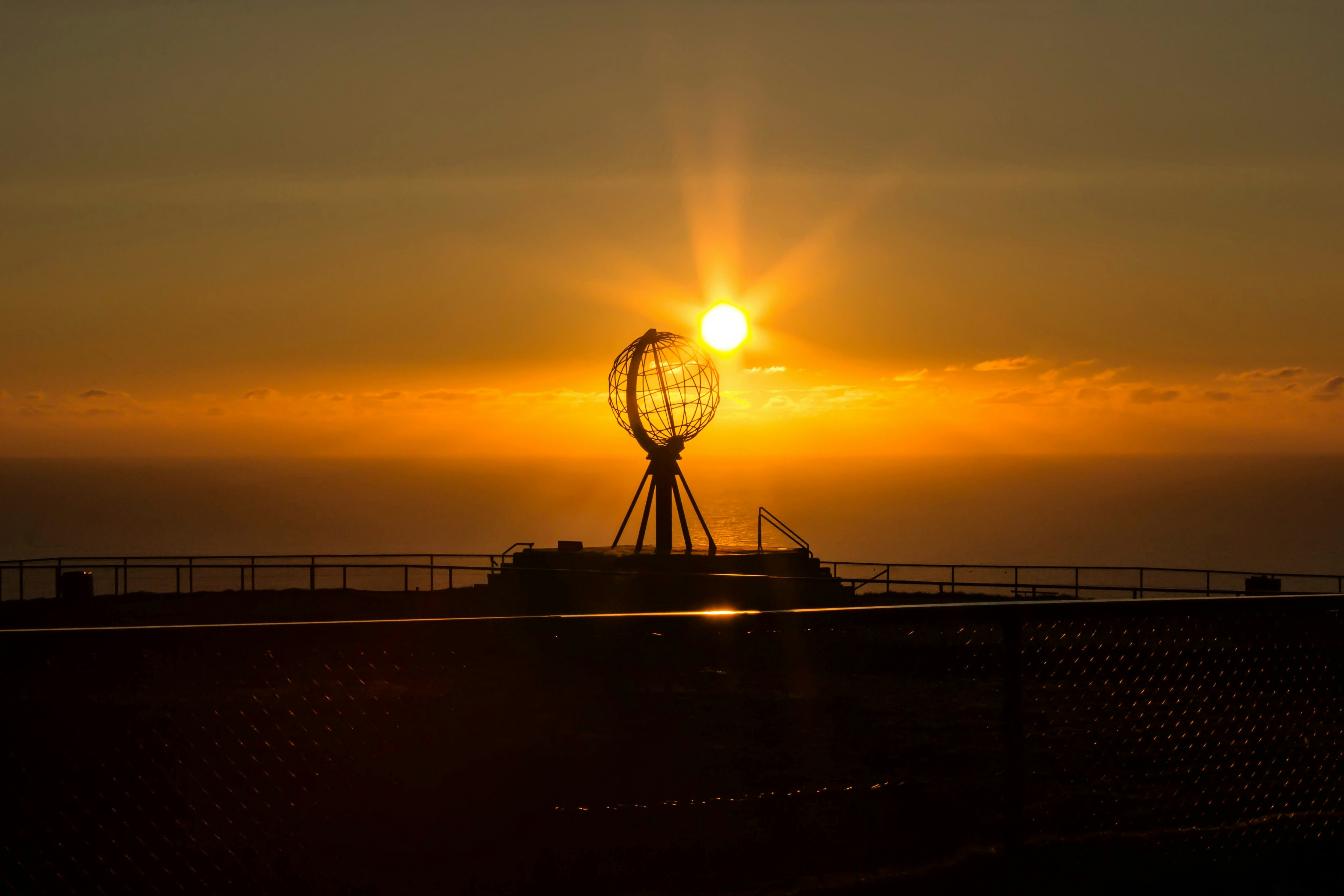 Globe monument silhouetted against a bright sunset over the ocean.