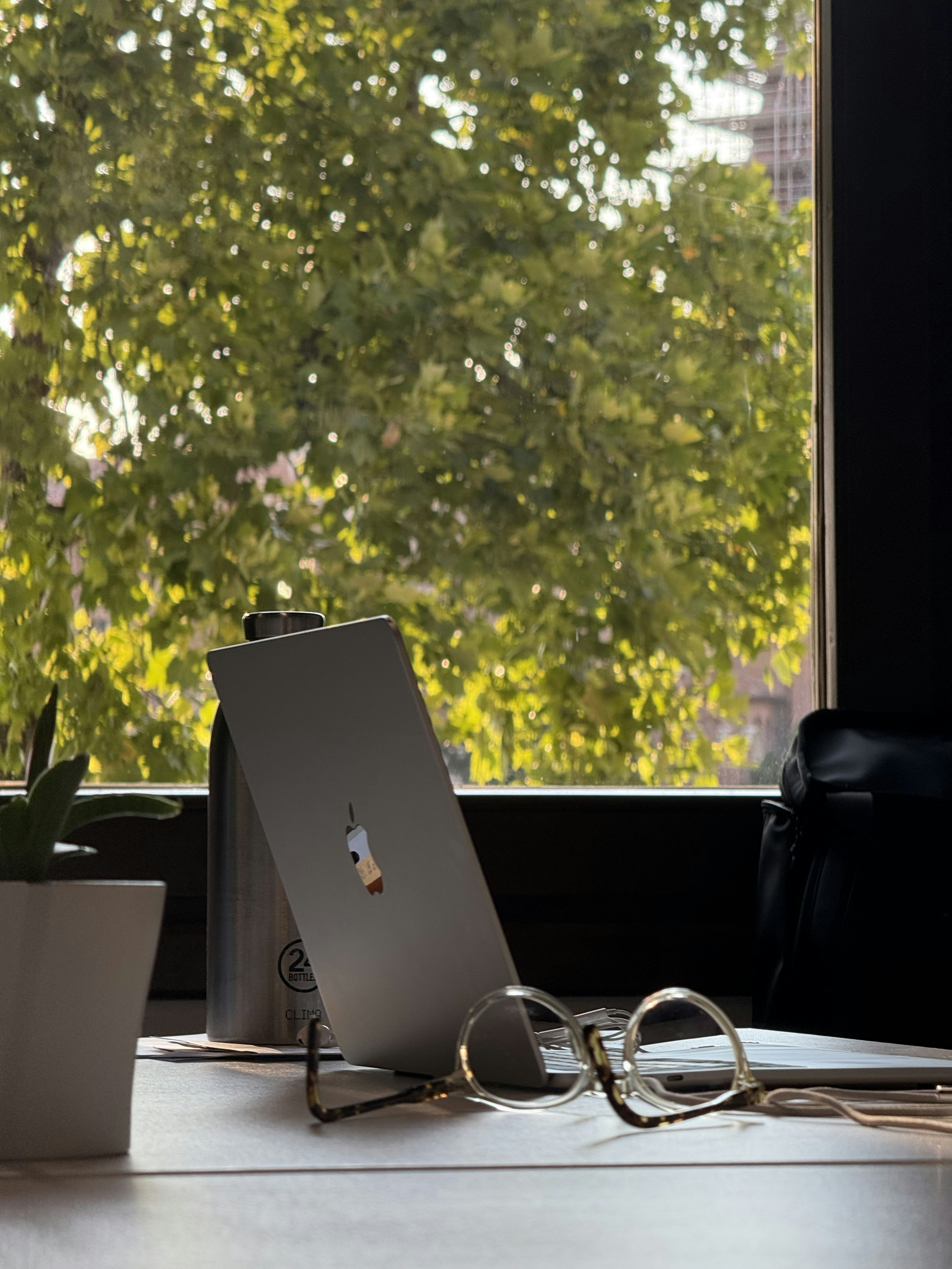 Laptop and glasses on desk with window view.