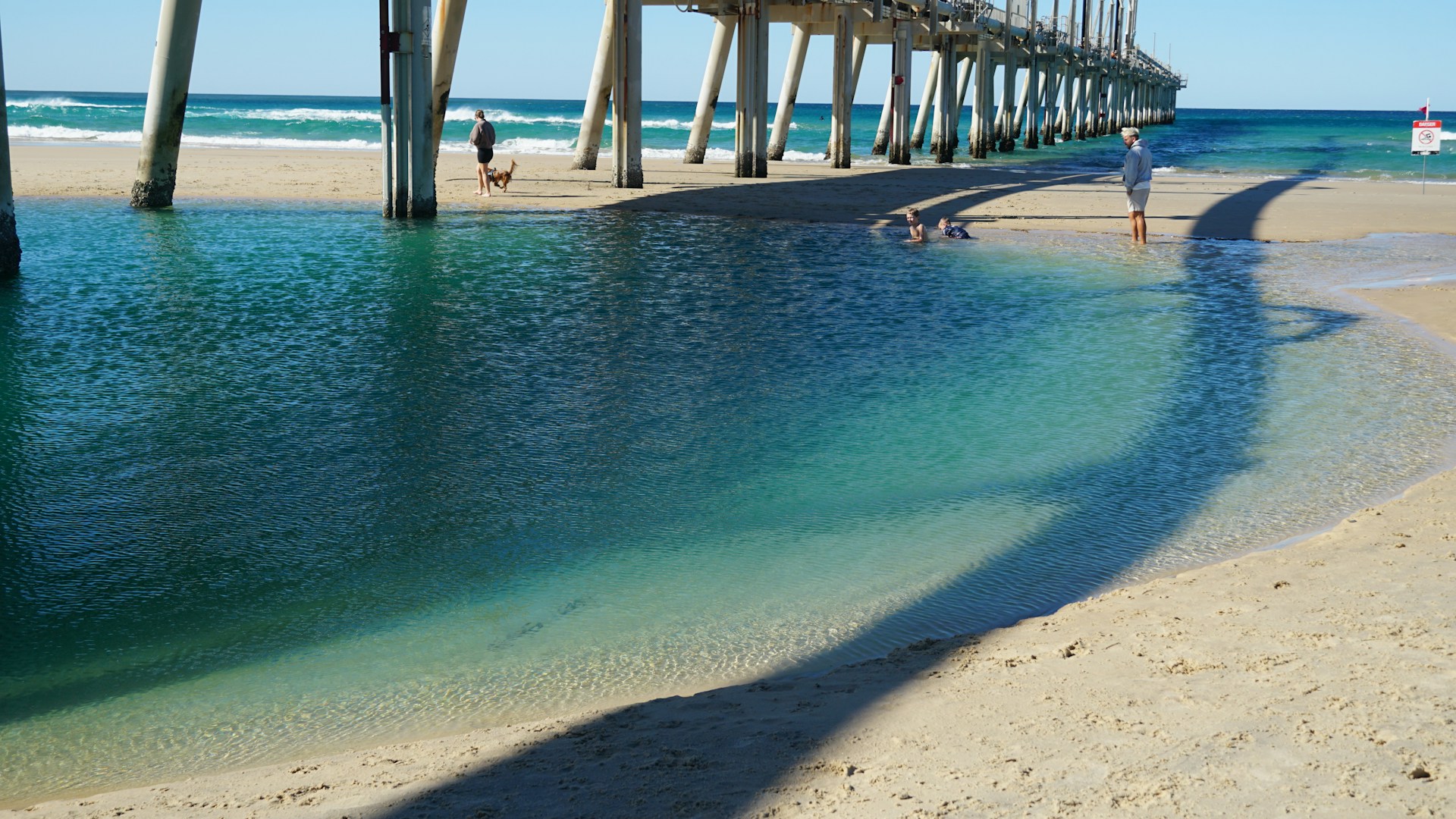 People walk on a sandy beach near a pier and ocean.