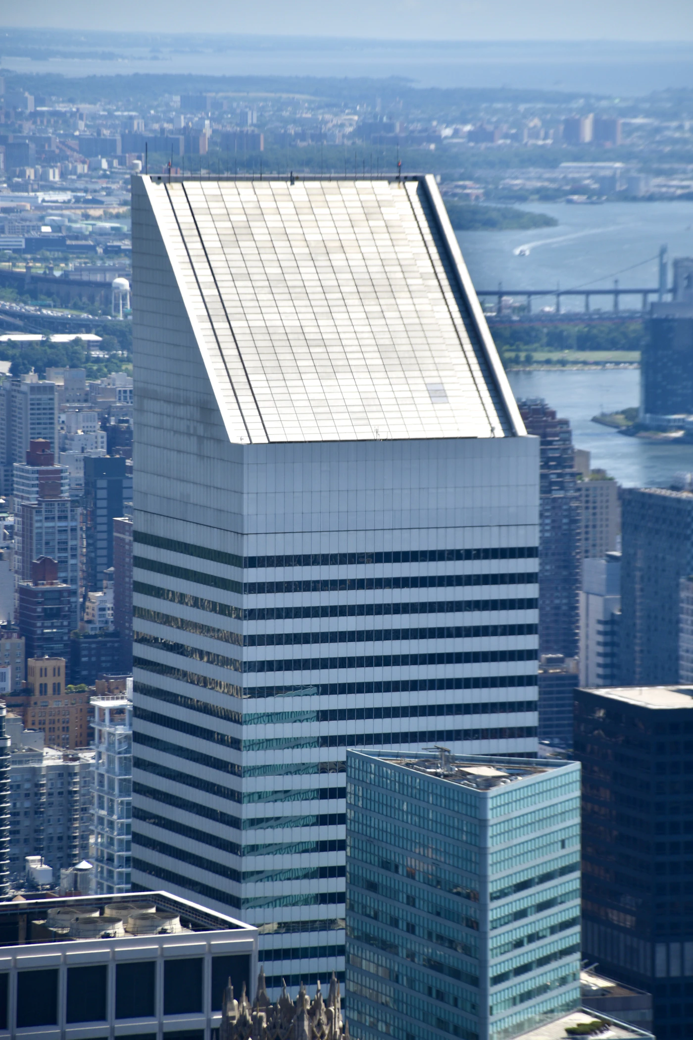 A skyscraper with a sharply slanted white roof, seen from above, with a river and a bridge behind it