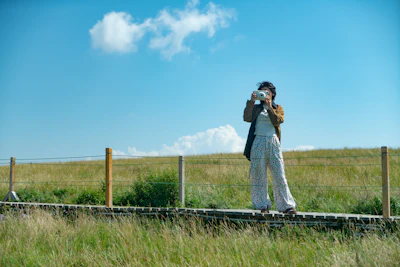 Woman with camera on wooden path in grassy field.