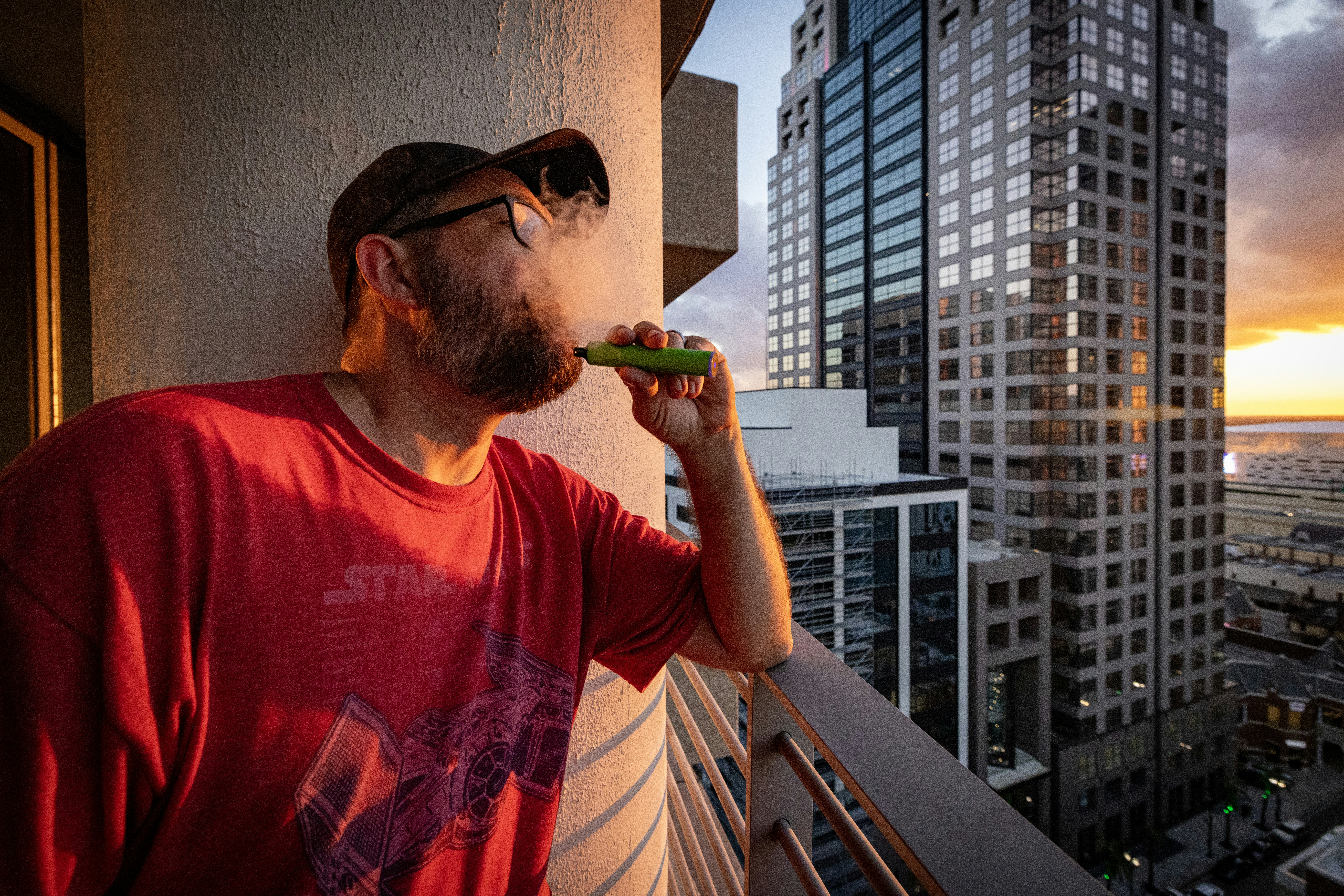 Man on balcony vaping with city skyline sunset.
