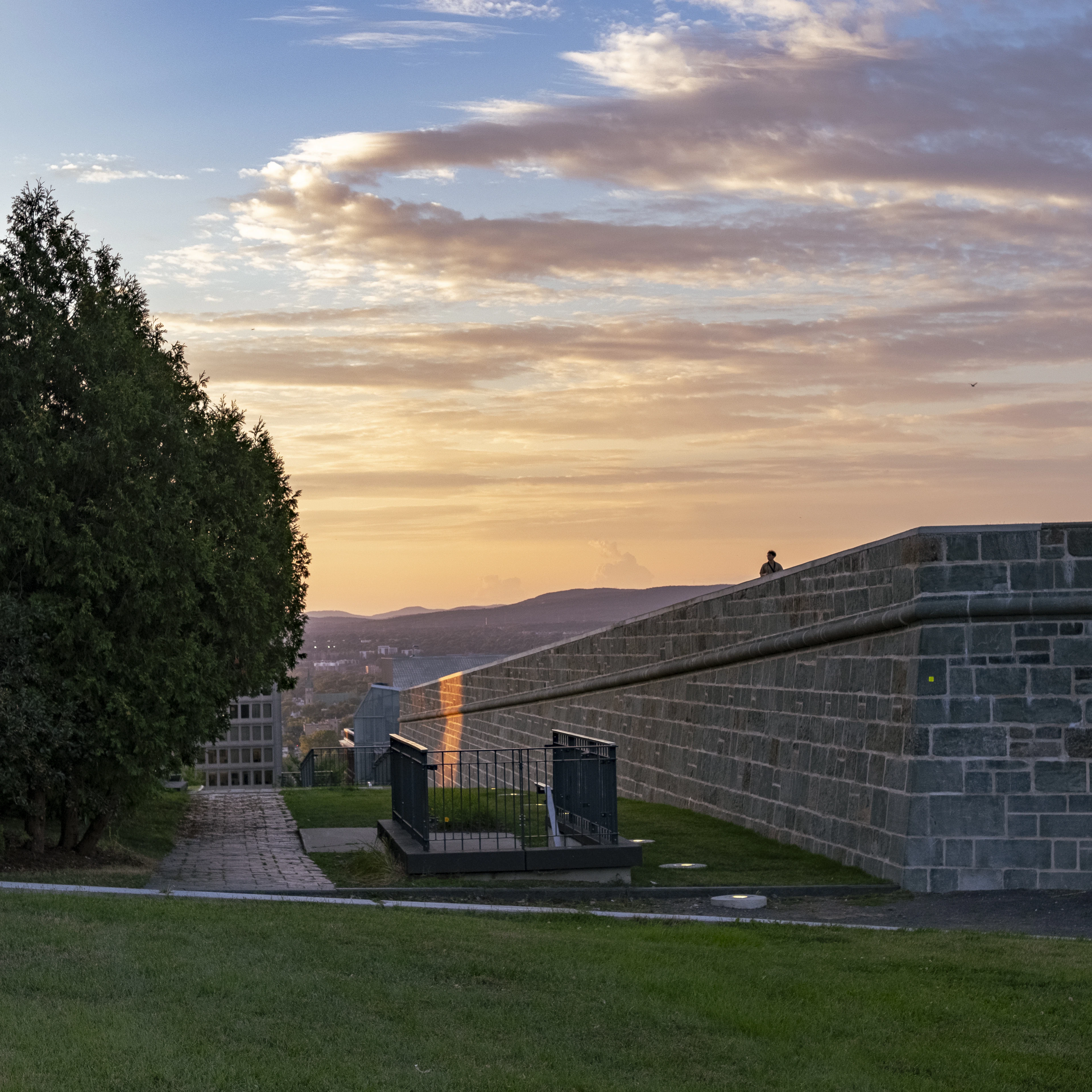 The stone ramparts of Old Quebec running down toward a gold and blue sunset over the distant hills, a single person sitting on top of the wall.