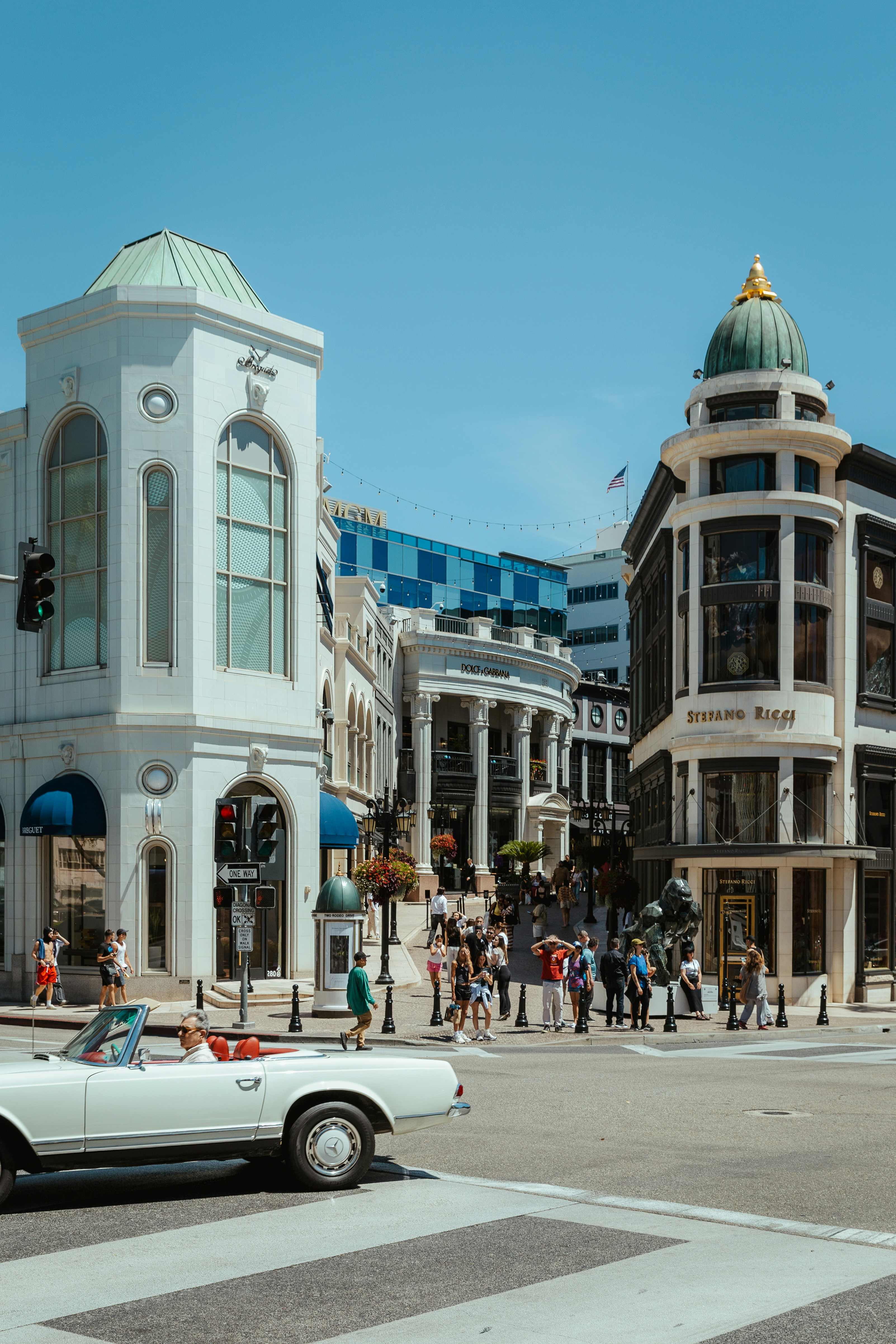 White convertible drives past luxury shops on sunny day.