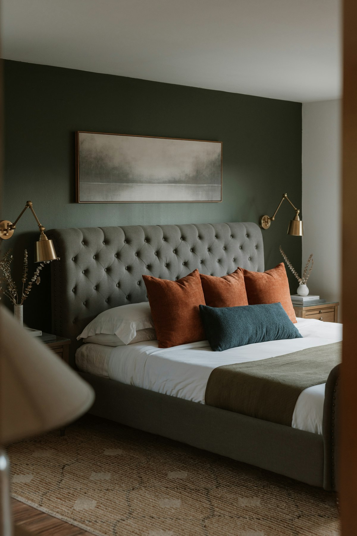 Cama de dos plazas con cabecera capitoné tapizada en tela gris, almohadones terracota y azul, apliques dorados y un cuadro sobre una pared verde oscuro
