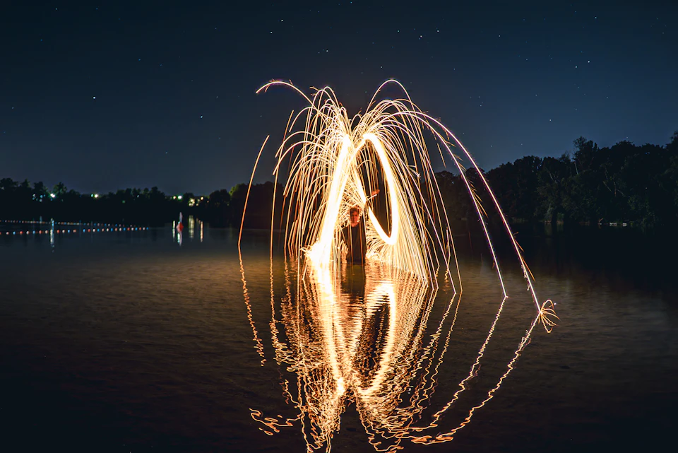 The image depicts a serene night scene by a lake, with a large group of fireflies illuminating the dark sky above. The fireflies are scattered across the frame, creating a mesmerizing display of light and shadow on the water's surface. In the center of the image, there is a person standing in front of a large body of water, their reflection mirrored perfectly in the calm waters below. The lake itself is surrounded by trees that add depth to the scene, with their silhouettes visible against the dark sky.