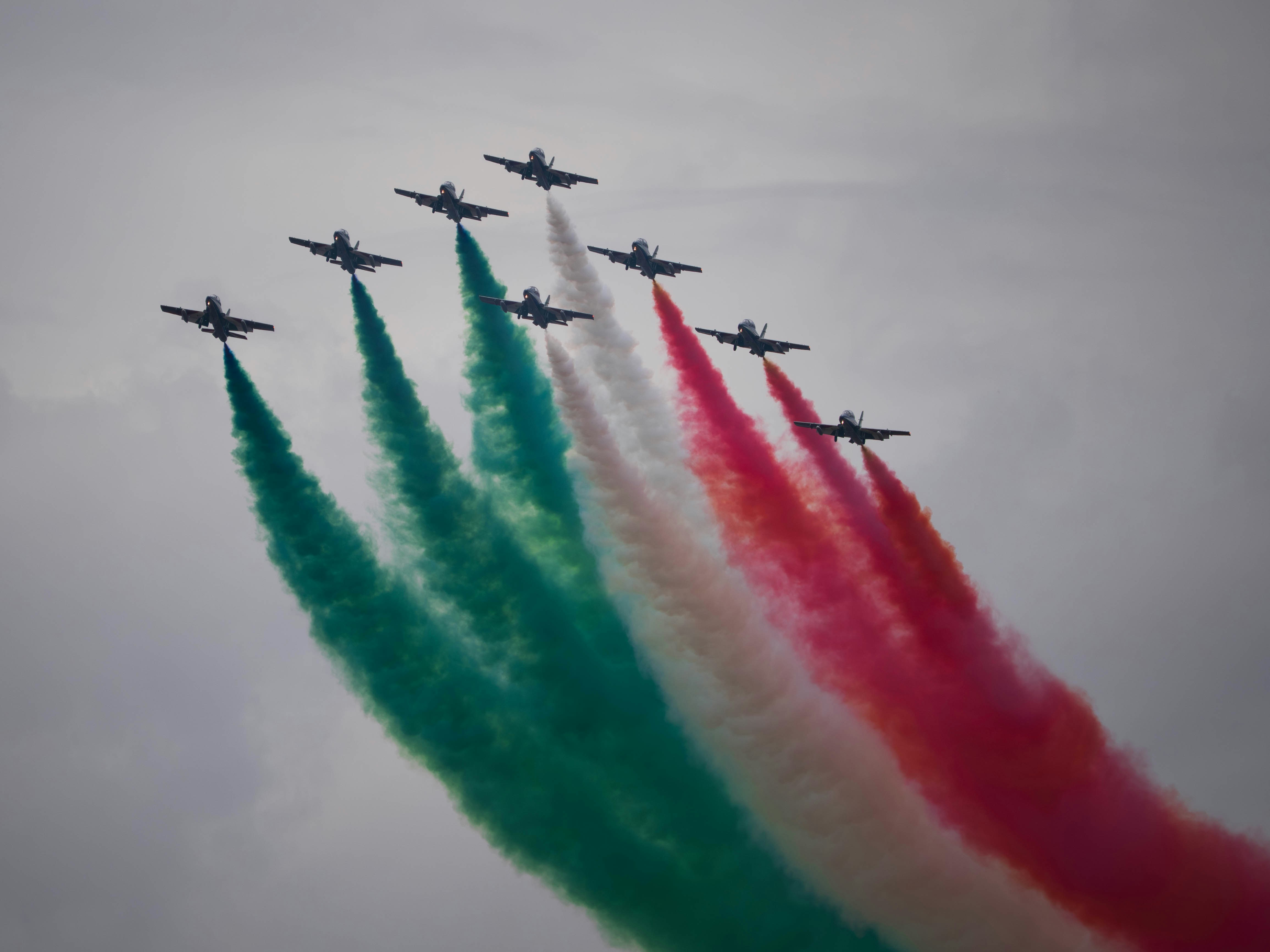 Nine fighter jets fly in formation leaving colored smoke trails.