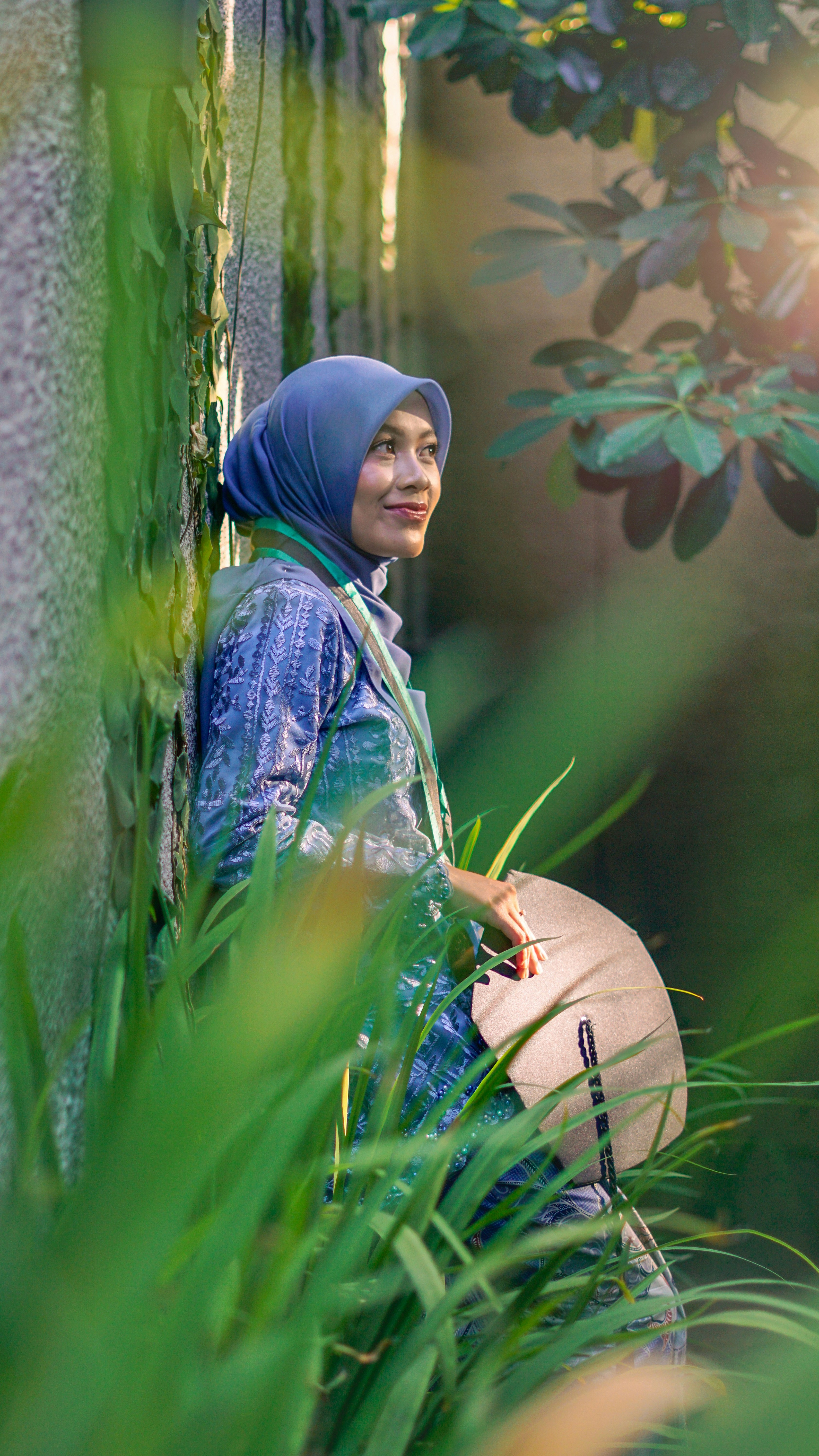 Woman in hijab leans against wall with fan.