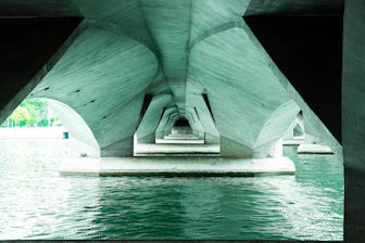 Underneath a modern concrete bridge with water below.