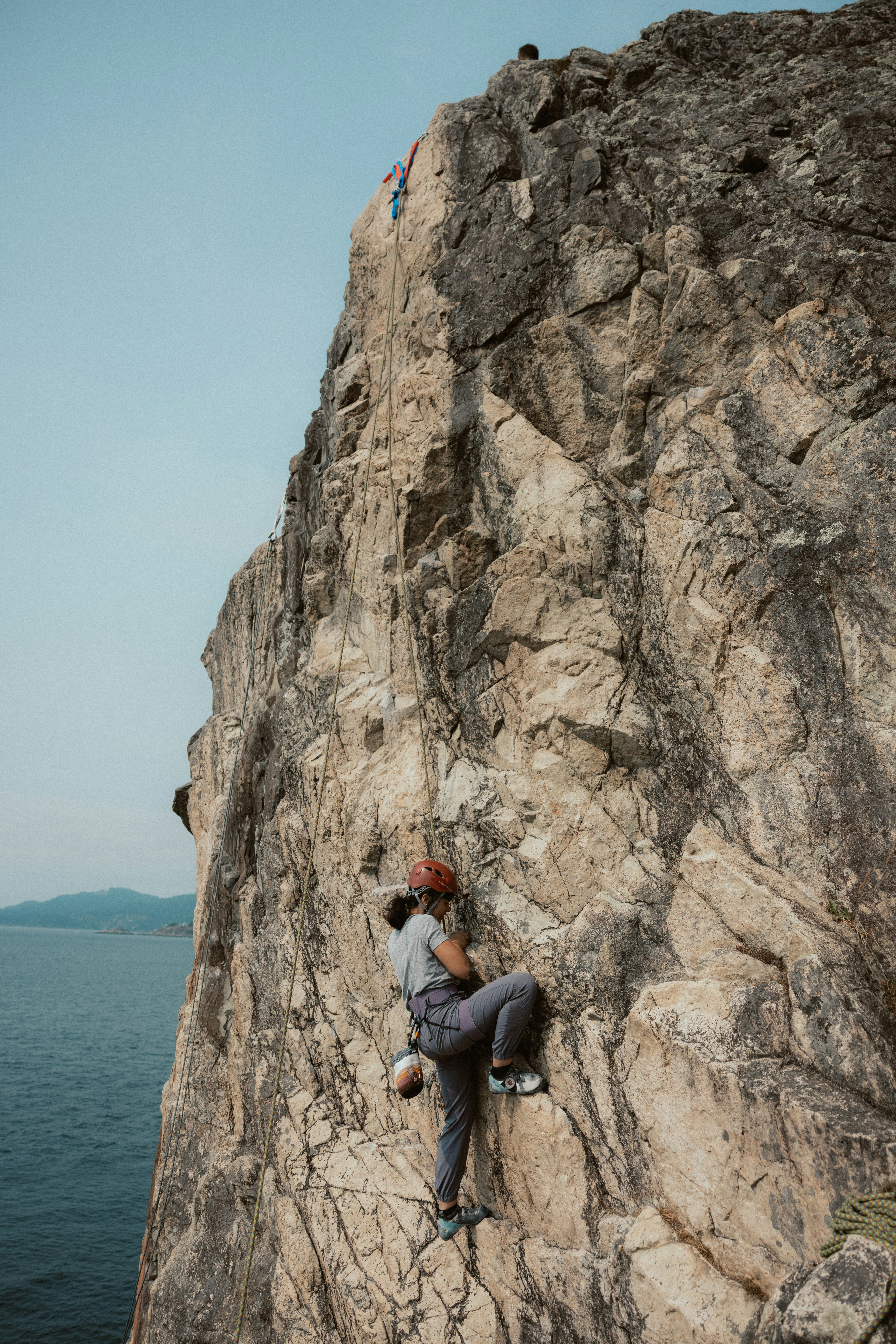 Rock climber ascends a steep cliff face near water.