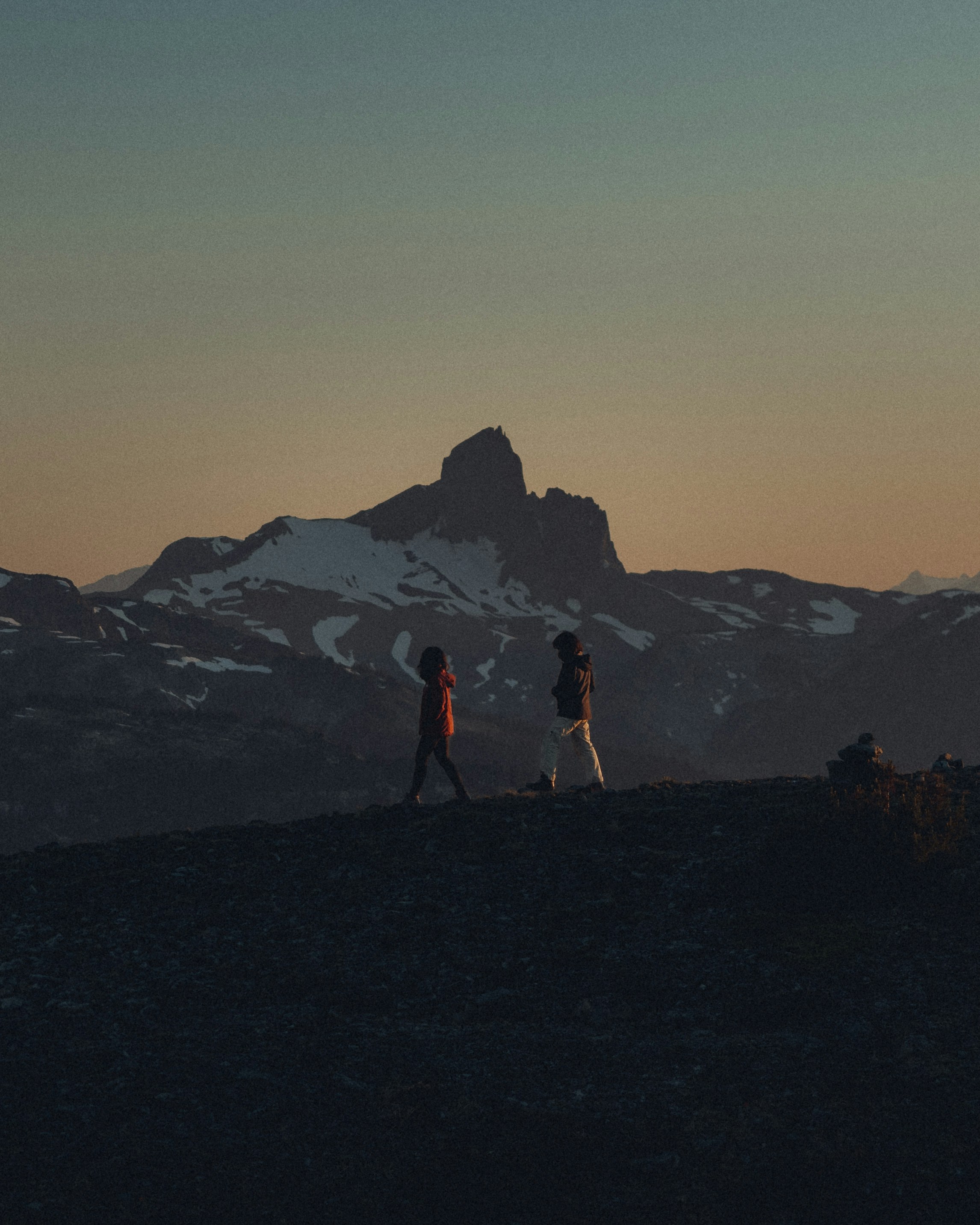 Two people walk on a ridge with mountains at sunset.