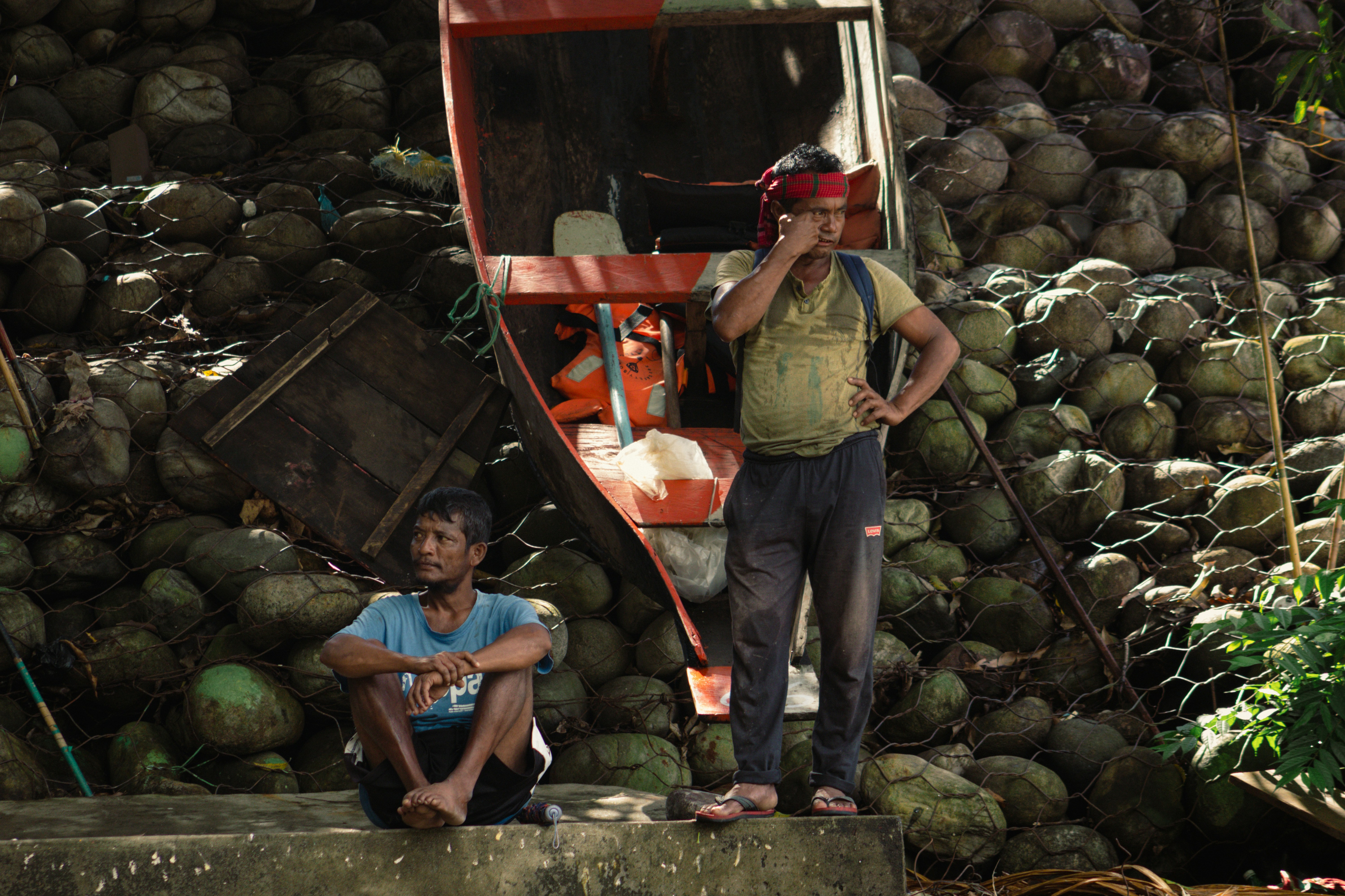 Dos hombres descansando cerca de un barco y una pared de cocos apilados.