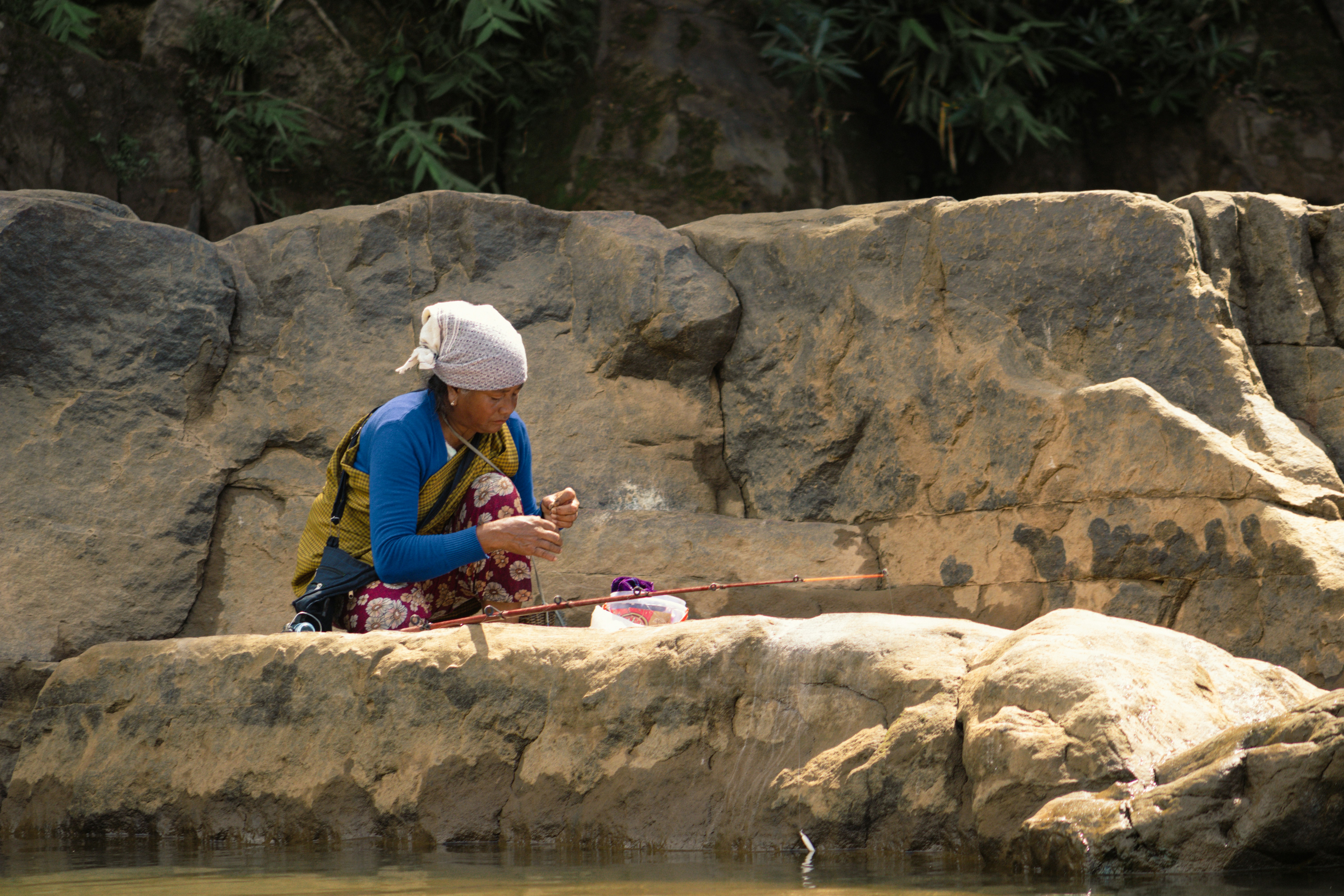 A woman in a headscarf fishing with a rod on large river rocks