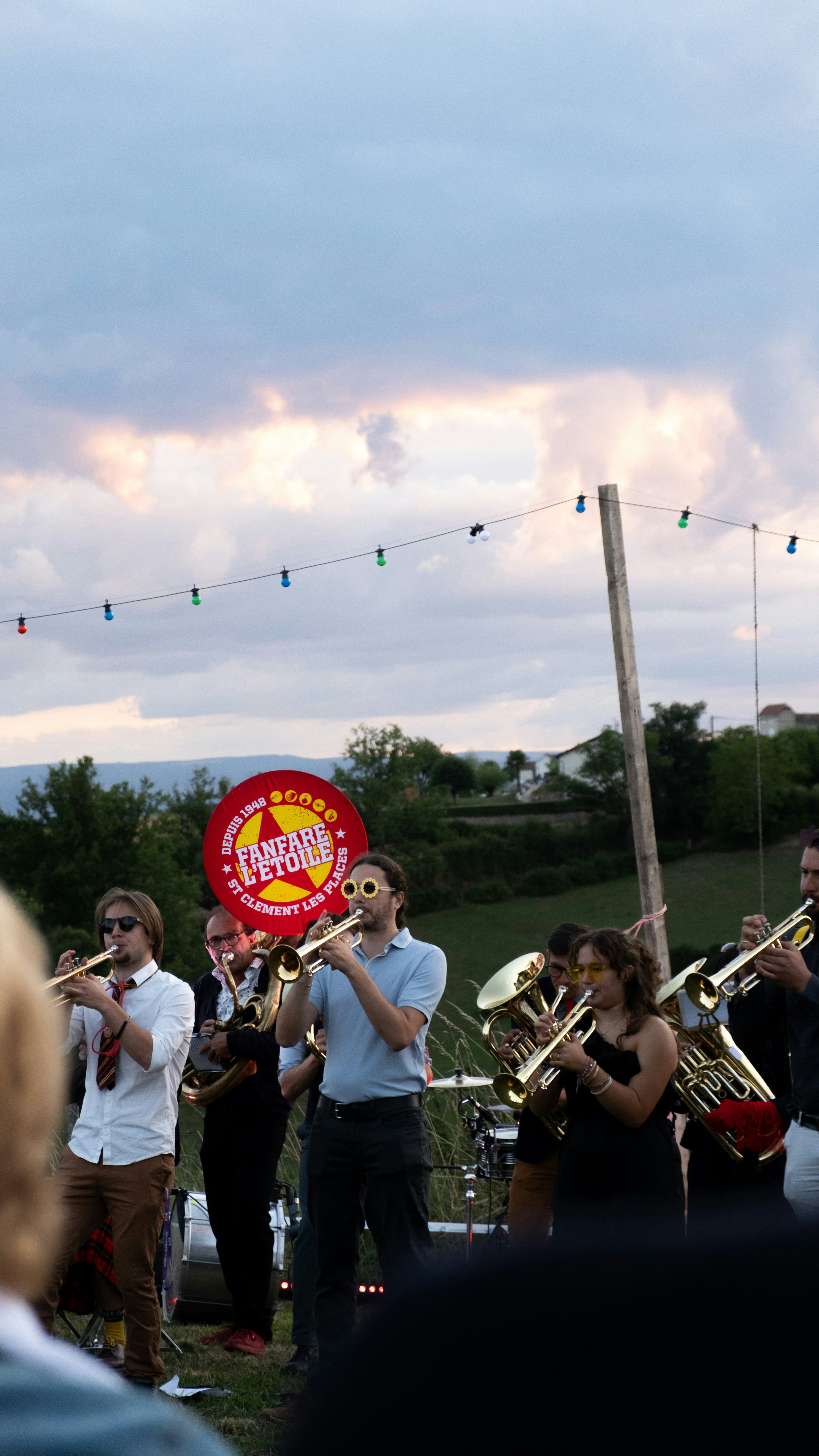 A brass band performs outdoors under string lights at dusk.