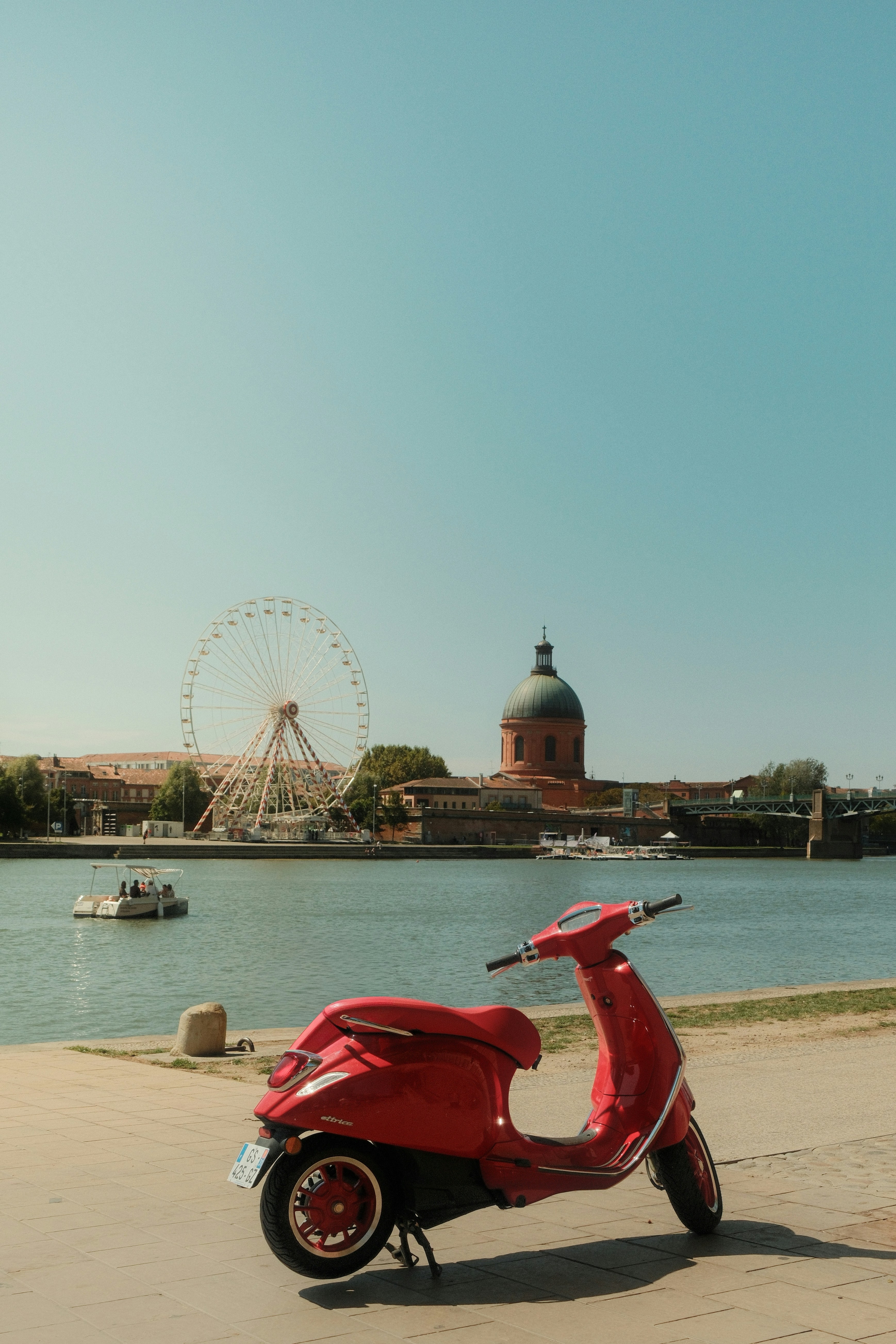 Scooter rouge garé au bord d’une rivière avec grande roue.