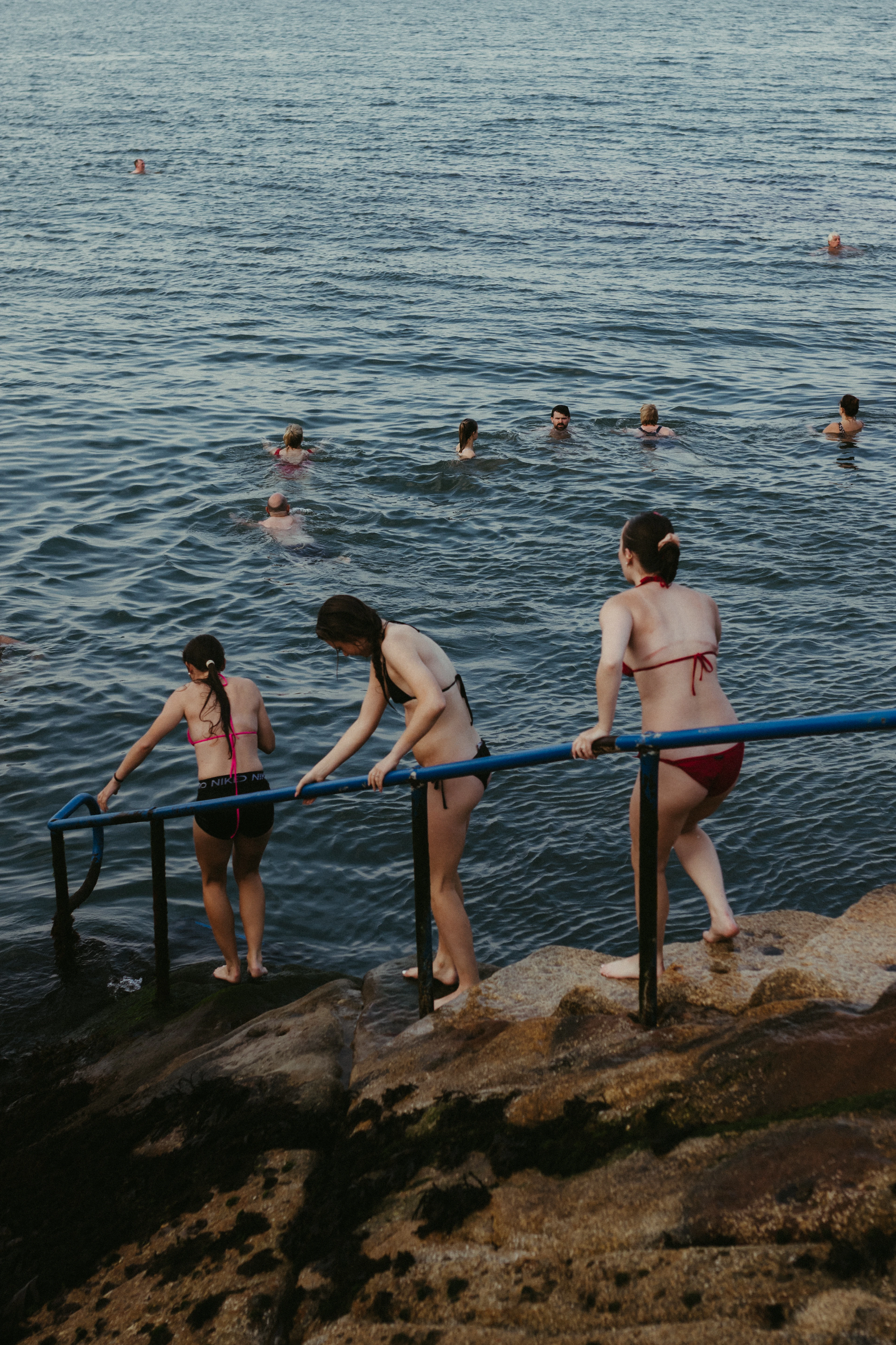 Women wade into the sea from a rocky shore at a Dublin swimming spot in evening light.