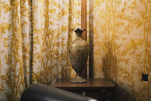 Ornate vase on a wooden table with patterned curtains.