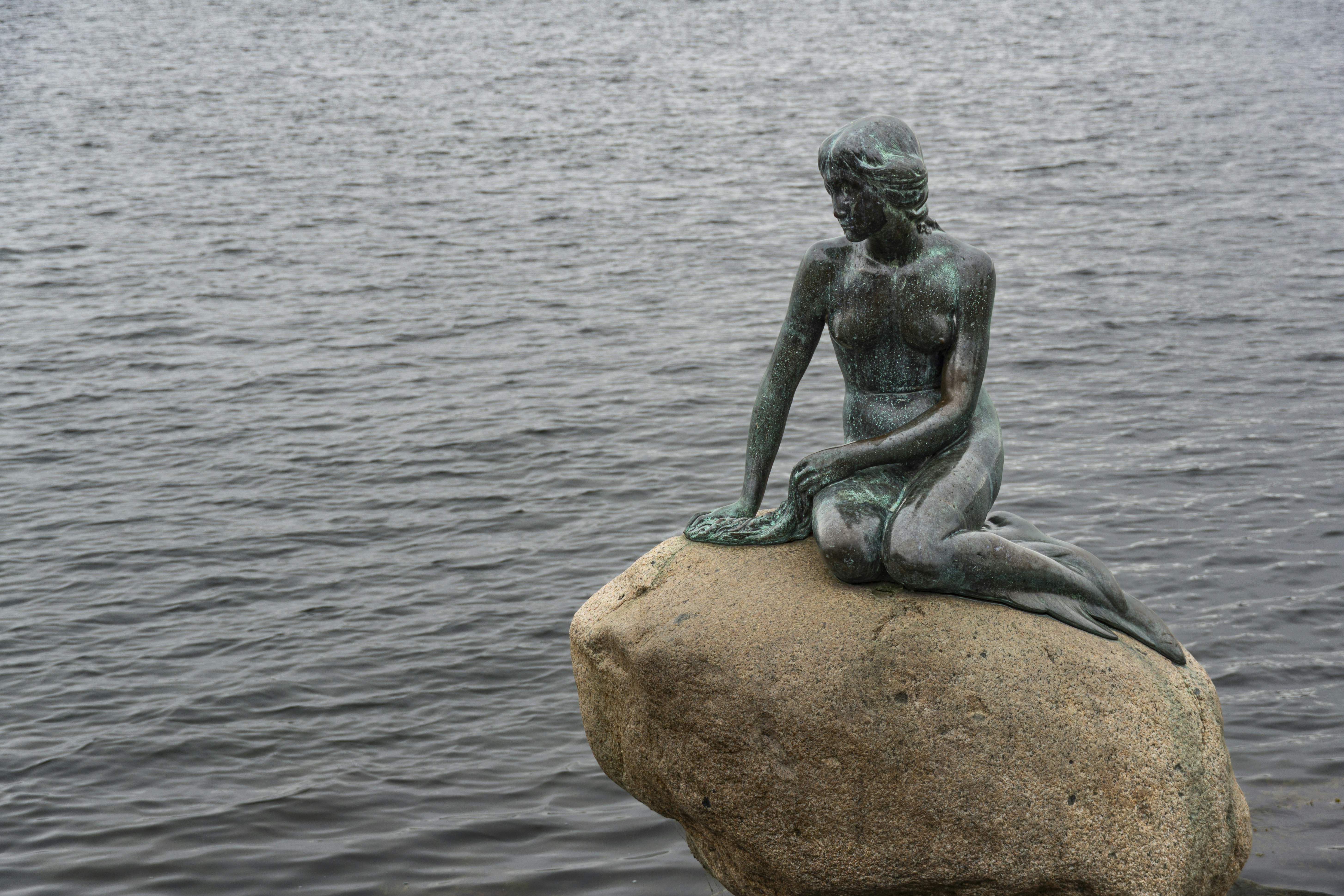 Die bronzene Meerjungfrauenstatue sitzt auf einem Felsen am Wasser.