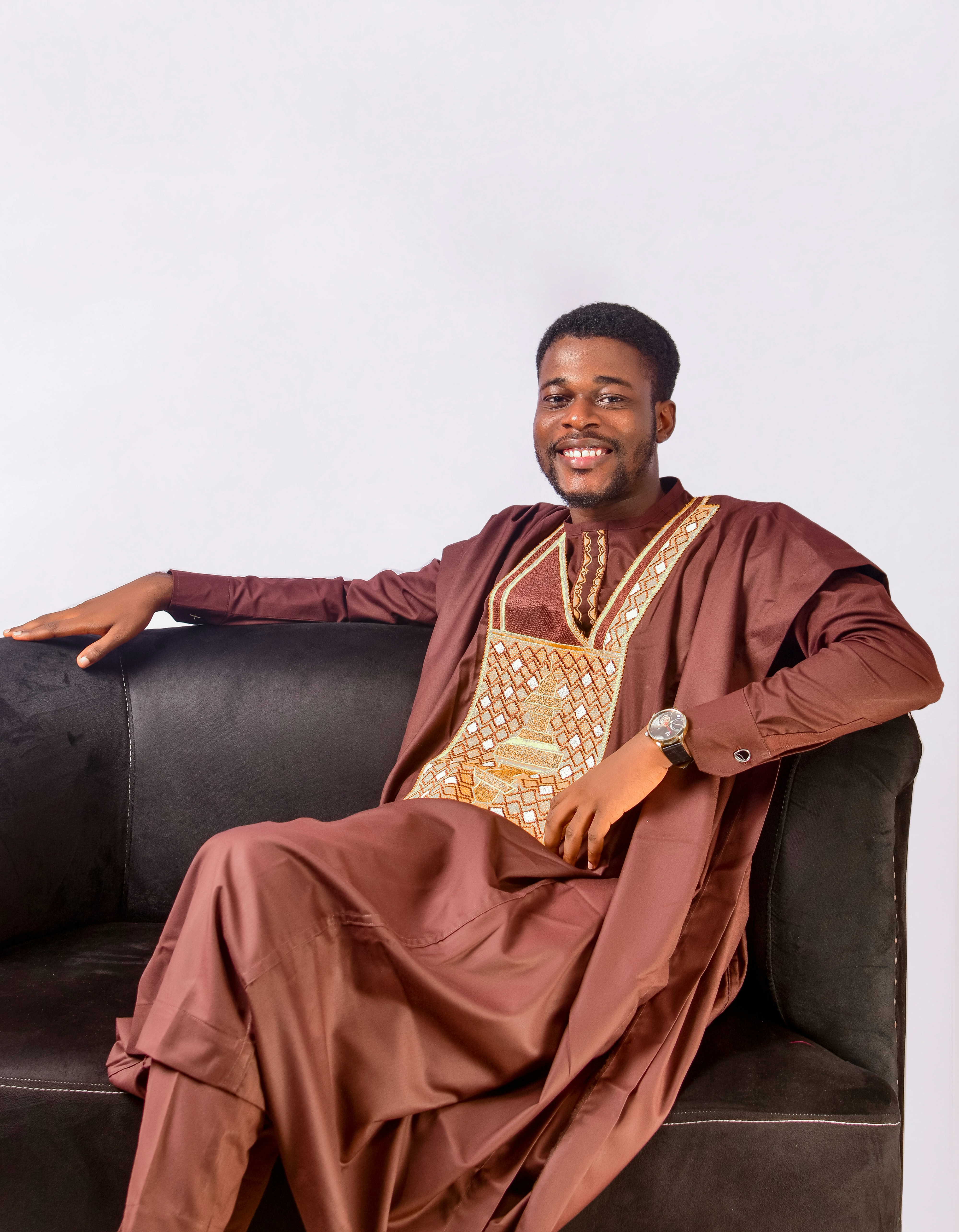 A man in brown traditional attire smiles on a couch.