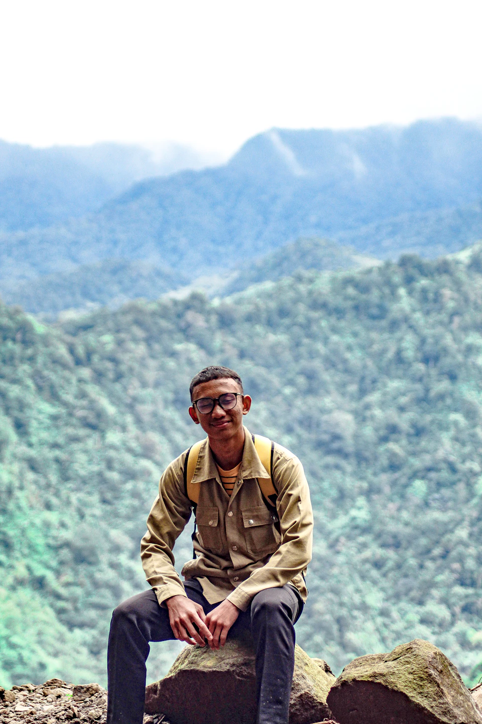In the image, a man is sitting on a large rock formation with his legs crossed and arms resting on his knees. He is wearing a beige shirt and glasses, giving him an intellectual appearance. The man has a backpack on his shoulders, suggesting he may be on a hike or exploring the outdoors. The background features lush green trees and rolling hills, indicating that they are in a mountainous region with dense vegetation.