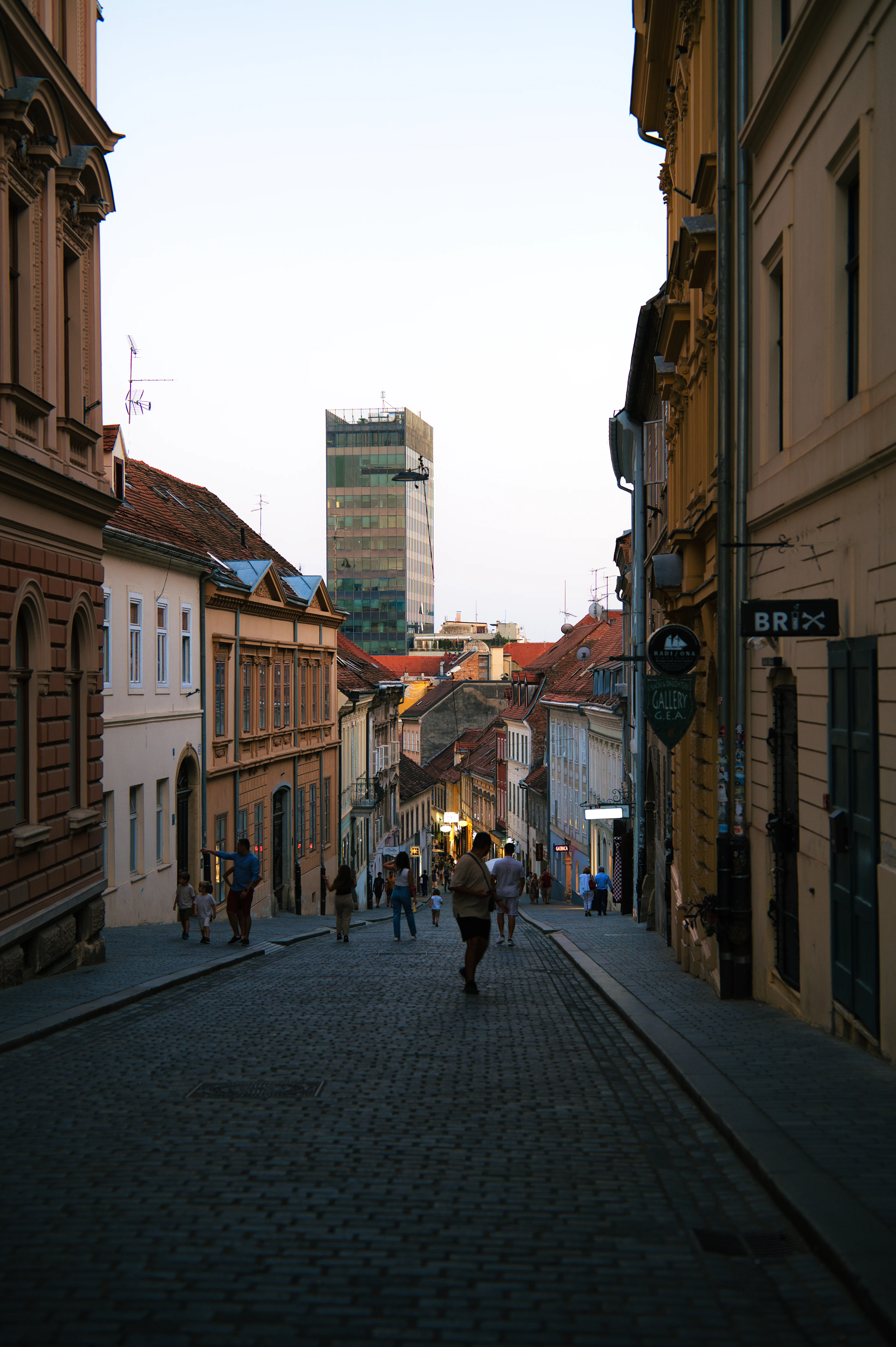 Pedestrians walk a cobblestone old-town street at dusk with a modern glass skyscraper rising behind the rooftops.