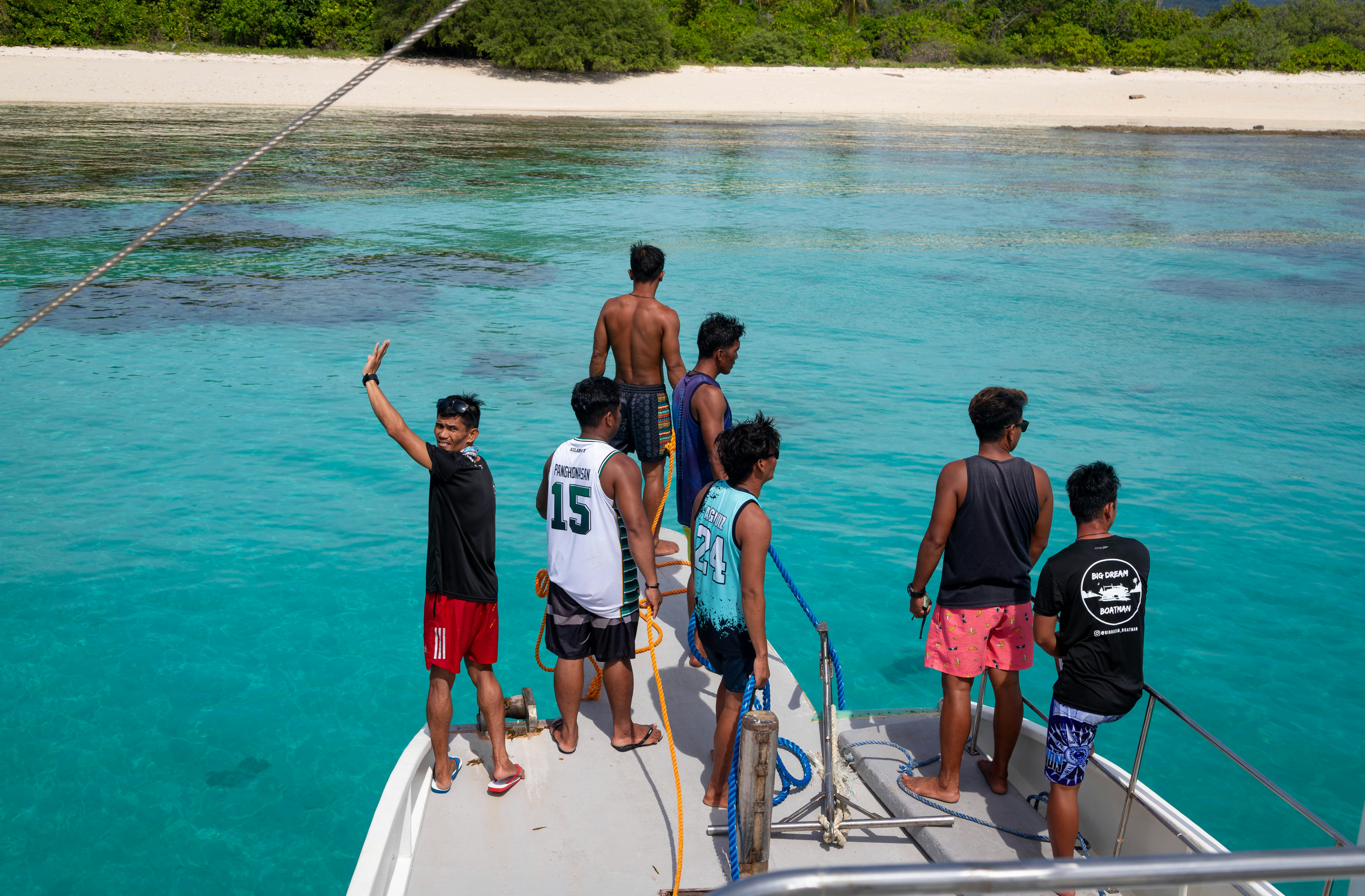A group of men stands at the bow of a boat positioned over clear turquoise water near a coral reef and a sandy beach. They are handling ropes and appear to be preparing to dock. The backdrop includes shallow water, green vegetation, and a tropical shoreline under sunny skies.