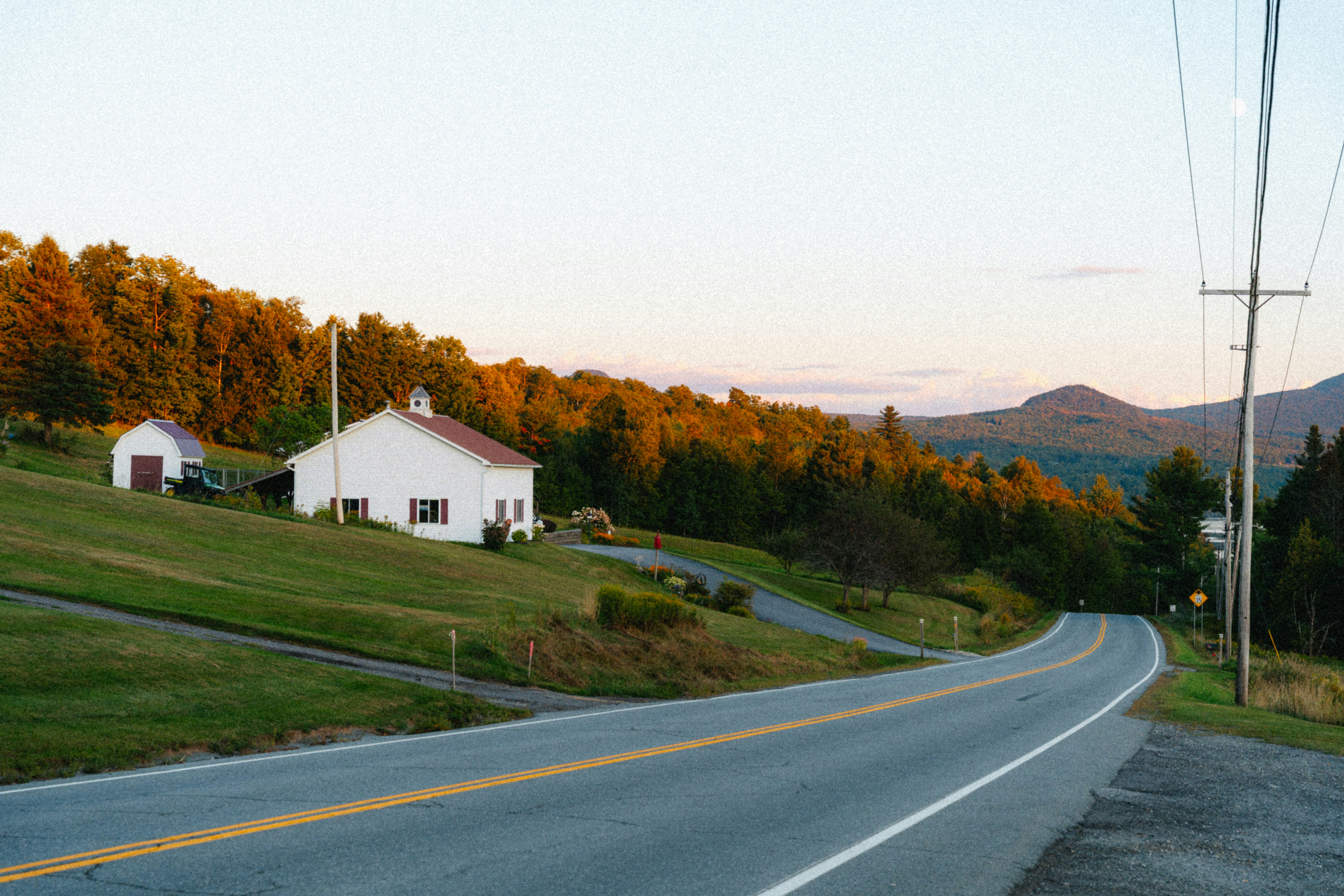 Rural road winds past white house and autumn trees.