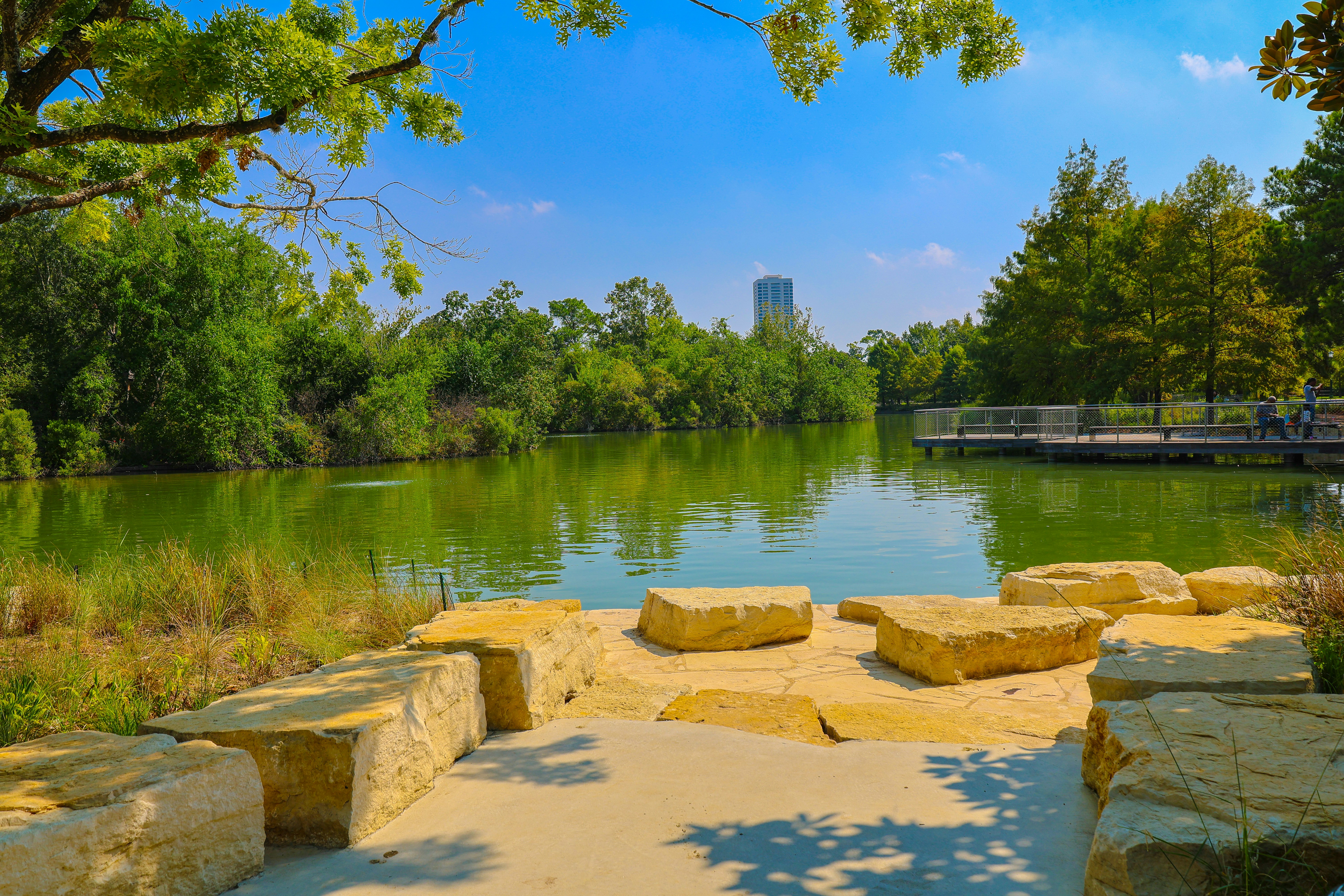 Sunny park with a lake and stone seating area.