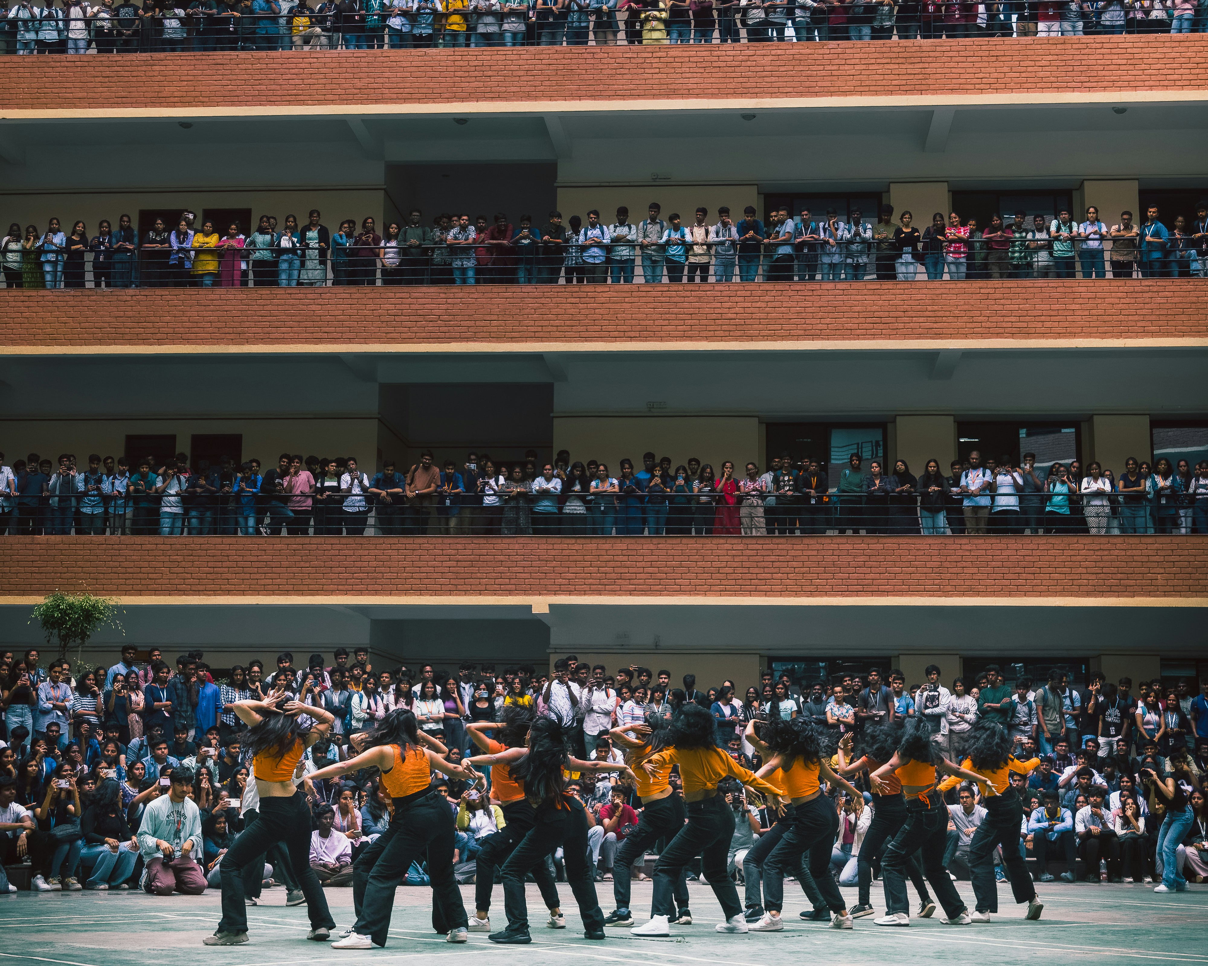 Dancers in orange tops perform for an audience in a courtyard.