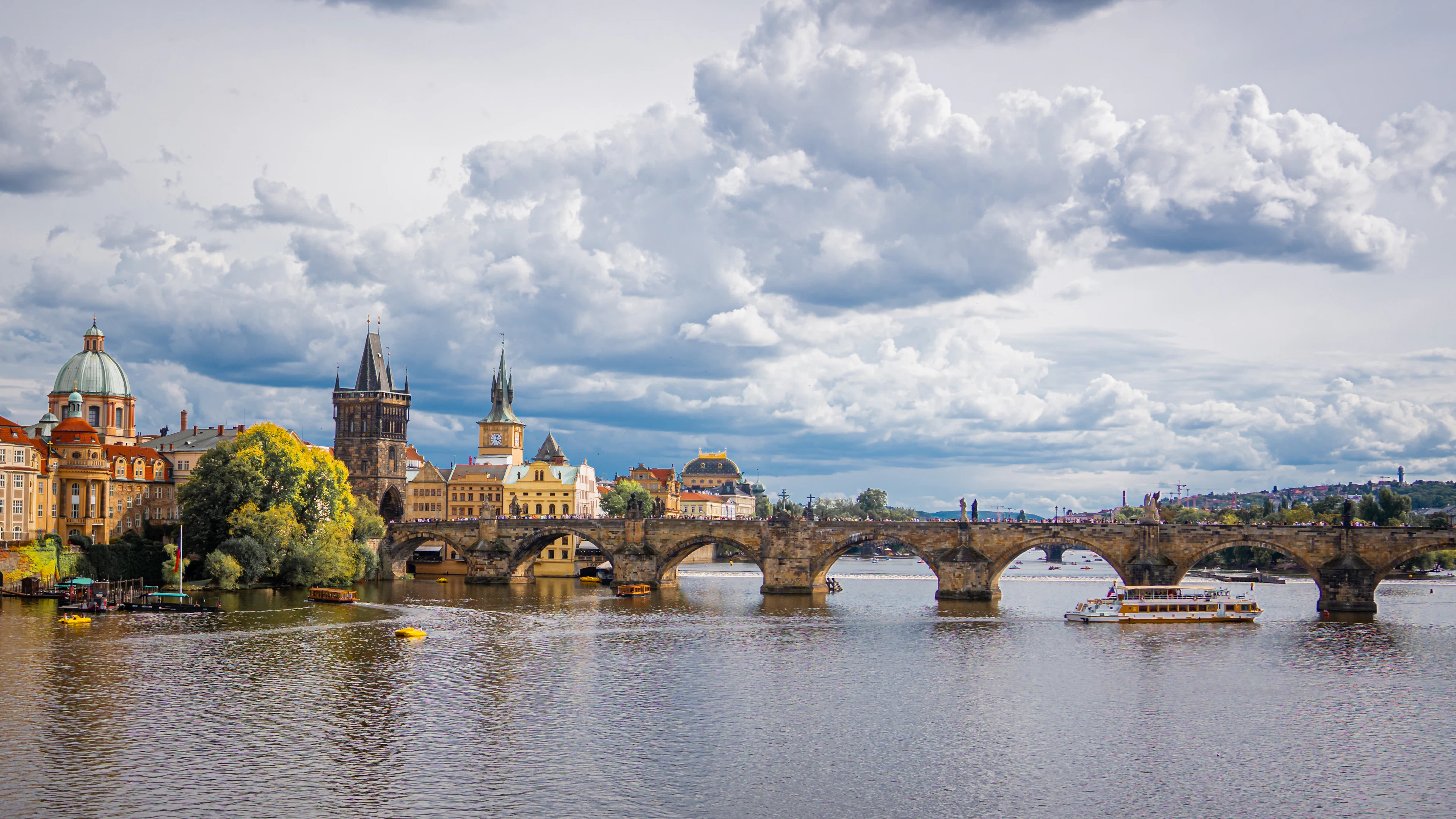 Charles Bridge spans the Vltava beneath dramatic clouds, with autumn-colored trees along the riverbank.