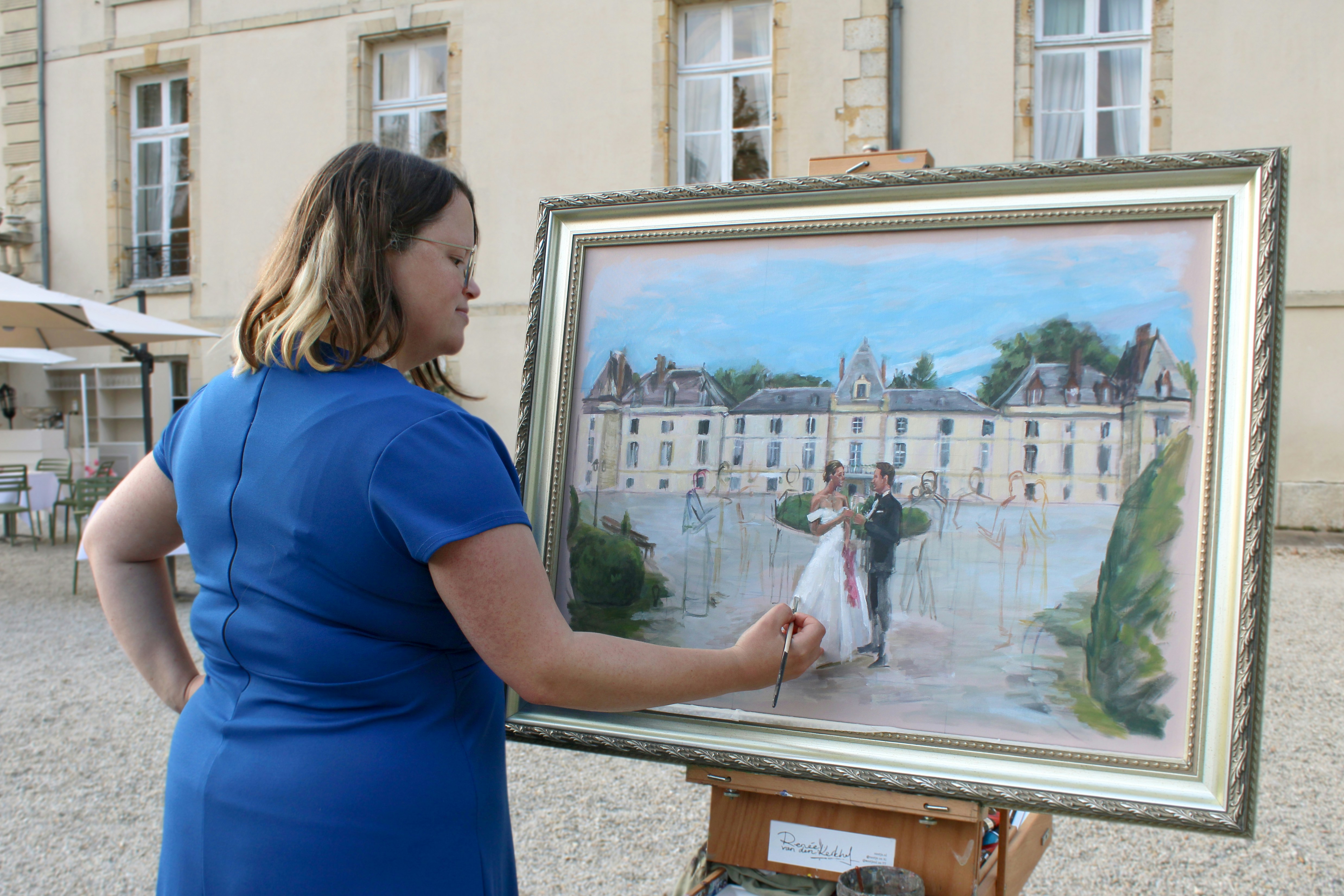 Live painting: female artist in blue dress at French castle - Château d'Aveny. Live wedding painting in a decorative frame, work in progress, The background and the wedding couple have a few details but the other people are still rough sketches. Live painter Renée looks into the camera, wears a blue maxi dress, painting an outdoor wedding scene with the couple in the center. Lovely, luxury 18th century castle. The artist is working with a paintbrush and a foldable French box easel. In the background we see part of the castle.
