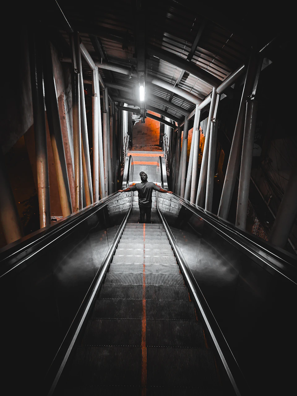 The image depicts a person standing on an escalator, facing away from the camera and looking down at the ground level. The escalator is black with orange stripes running along its length, providing a stark contrast against the otherwise dark surroundings. In the background, there are several metal poles that add to the industrial atmosphere of the scene.