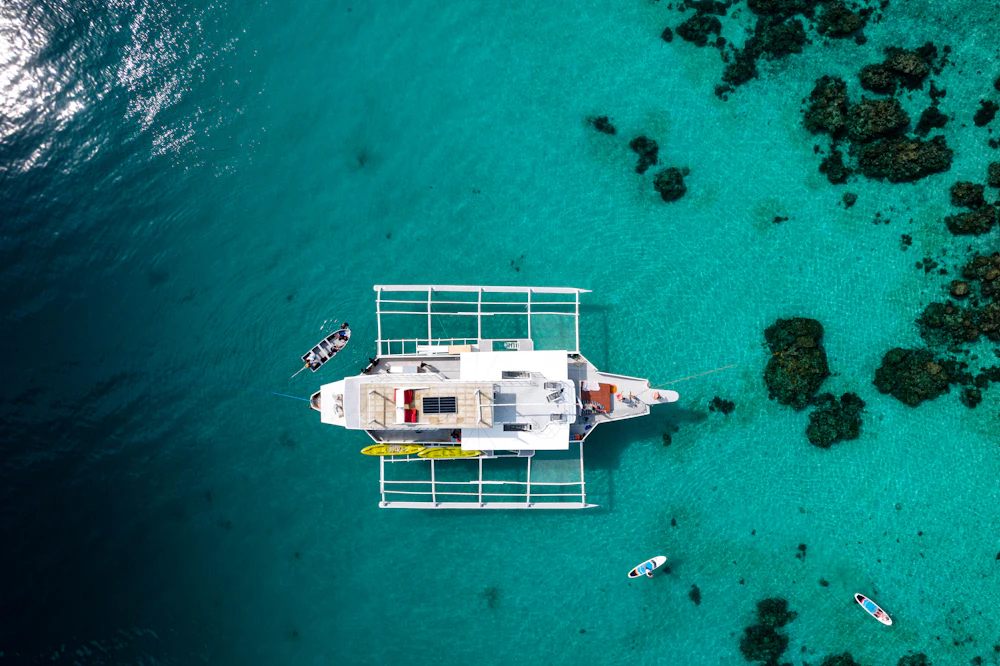A sailing catamaran seen from directly above, both hulls and the trampoline foredeck clear, lying over pale turquoise shallows.