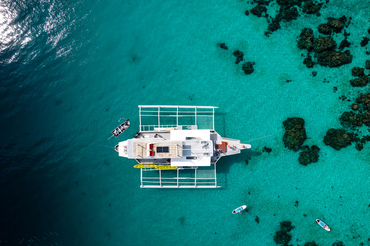 A sailing catamaran seen from directly above, both hulls and the trampoline foredeck clear, lying over pale turquoise shallows.