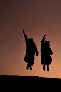 Two graduates in caps and gowns jumping at sunset.