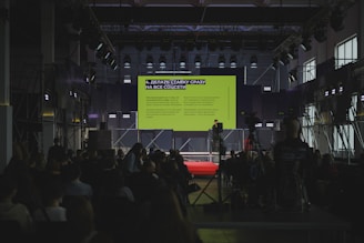 A speaker presents on a large stage with a screen.