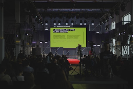 A speaker presents on a large stage with a screen.