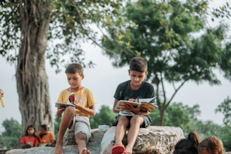Two boys reading books outdoors under a tree.