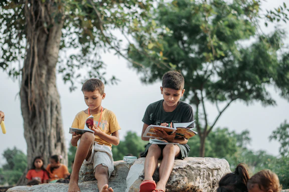 Two boys reading books outdoors under a tree.