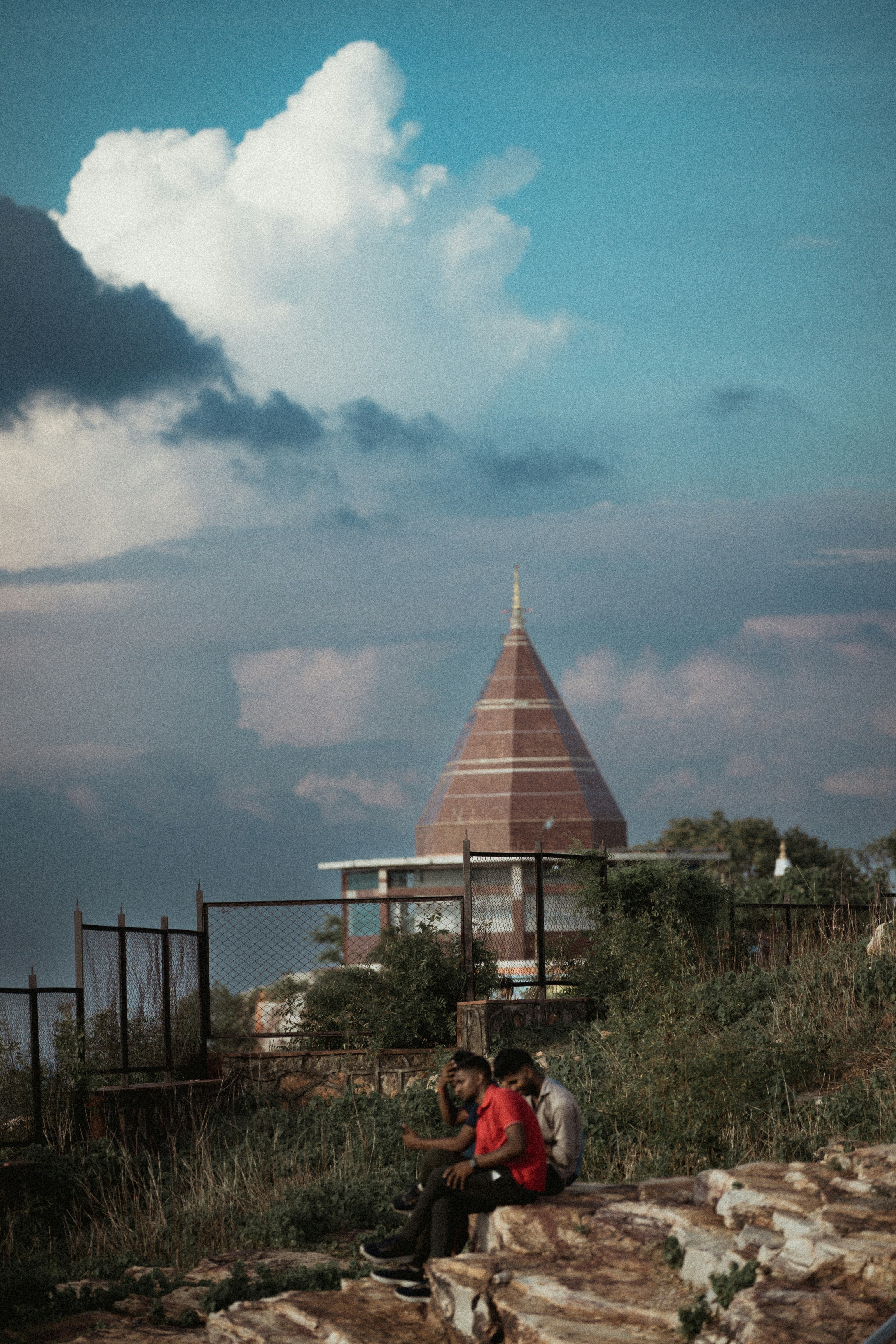 Two men sit near a conical building under clouds.