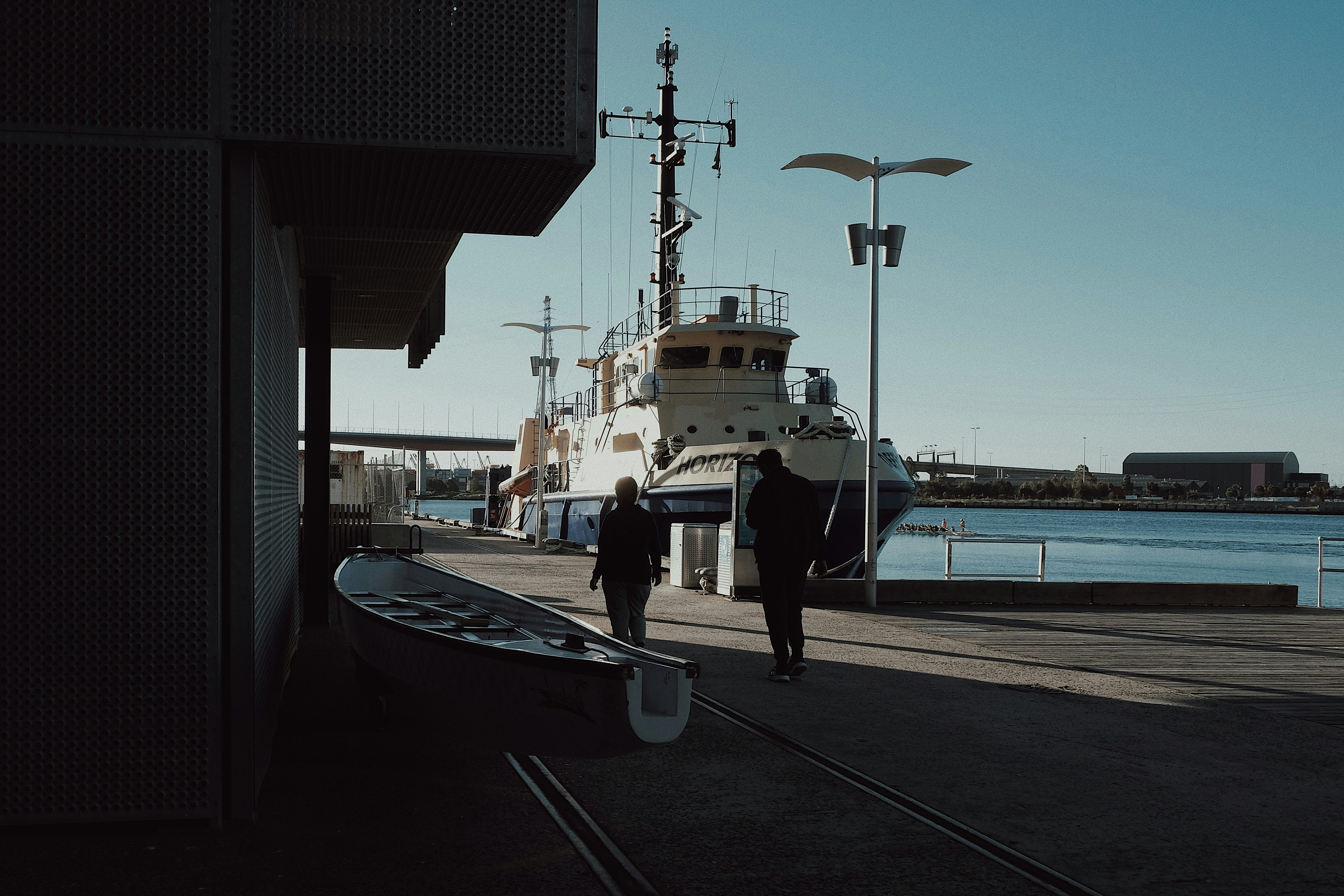 Two people walk past a boat near a docked ship.