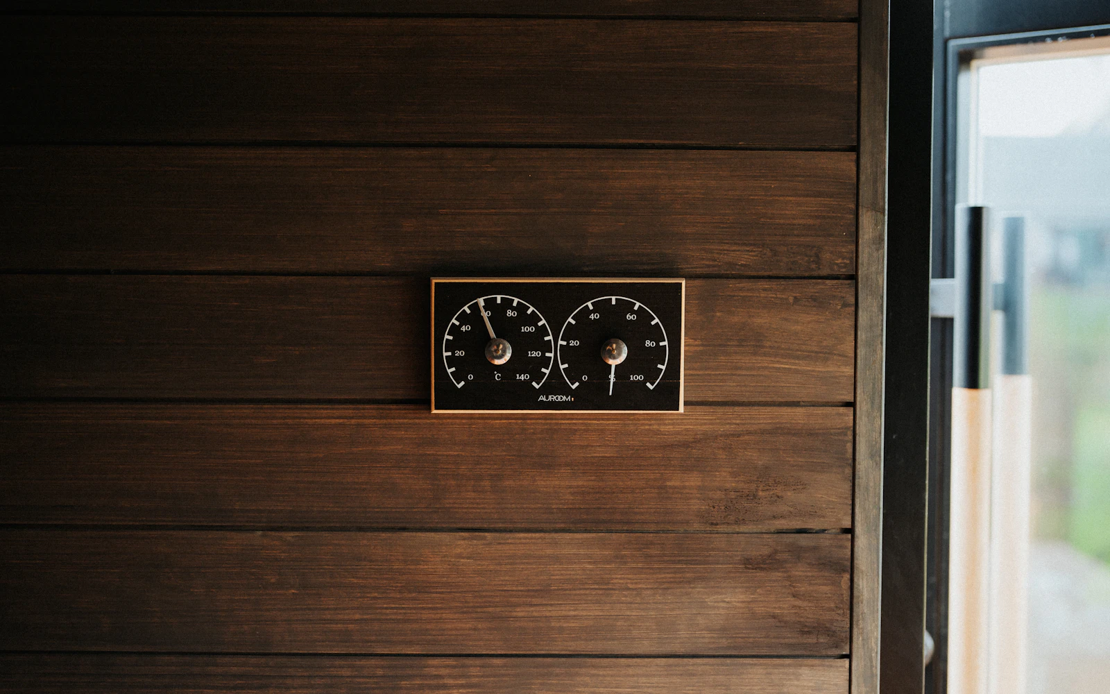 Paired analogue dials set in a wooden frame on a plank wall beside a window