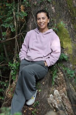 Woman in hoodie leans against large tree outdoors