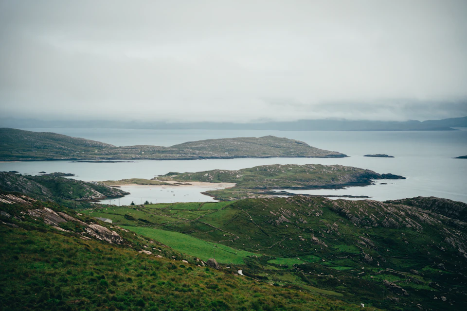 The image depicts a serene landscape with a large body of water, possibly an ocean or sea, nestled between two small islands. The island on the left is covered in lush green grass and dotted with trees, while the island on the right has rocky terrain and sparse vegetation. The sky above is overcast, casting a soft light over the scene. The perspective of the image suggests it was taken from a high vantage point, possibly from an airplane or helicopter, offering a bird's eye view of this picturesque landscape.