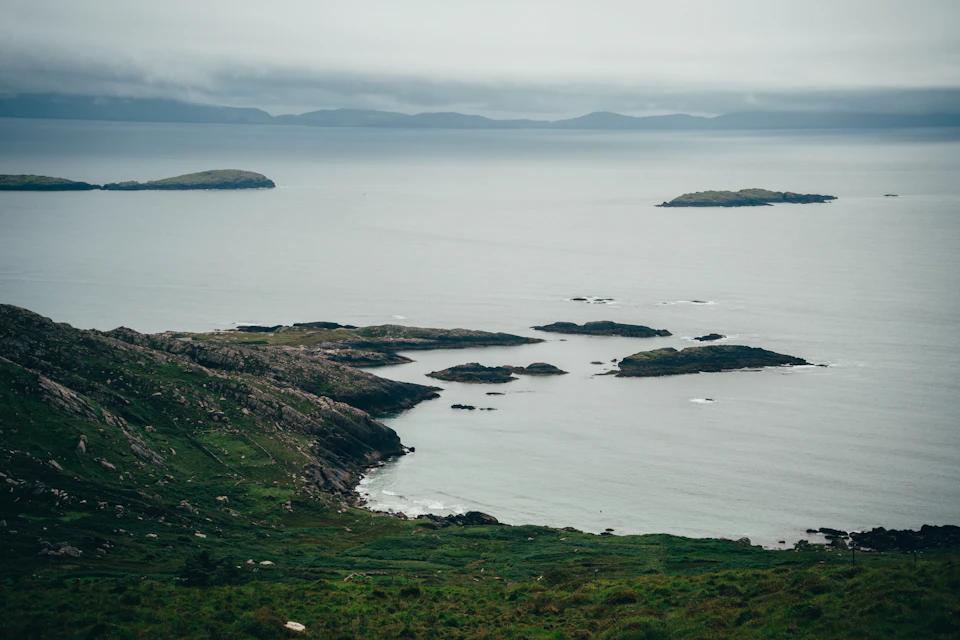The image depicts a serene coastal landscape with a rocky cliff overlooking a calm, light blue sea dotted with small islands scattered throughout. The sky above is overcast, casting a soft, diffused light over the scene. The perspective from the top of the cliff offers a panoramic view of the water and the surrounding land, creating a sense of tranquility and solitude.

The rocky cliff itself is rugged and uneven, with patches of greenery interspersed among the rocks. The sea below mirrors the sky above in its light blue hue, adding to the overall calmness of the scene. The small islands scattered across the water are mostly submerged, suggesting that they may be part of a larger body of water or simply isolated pieces of land.