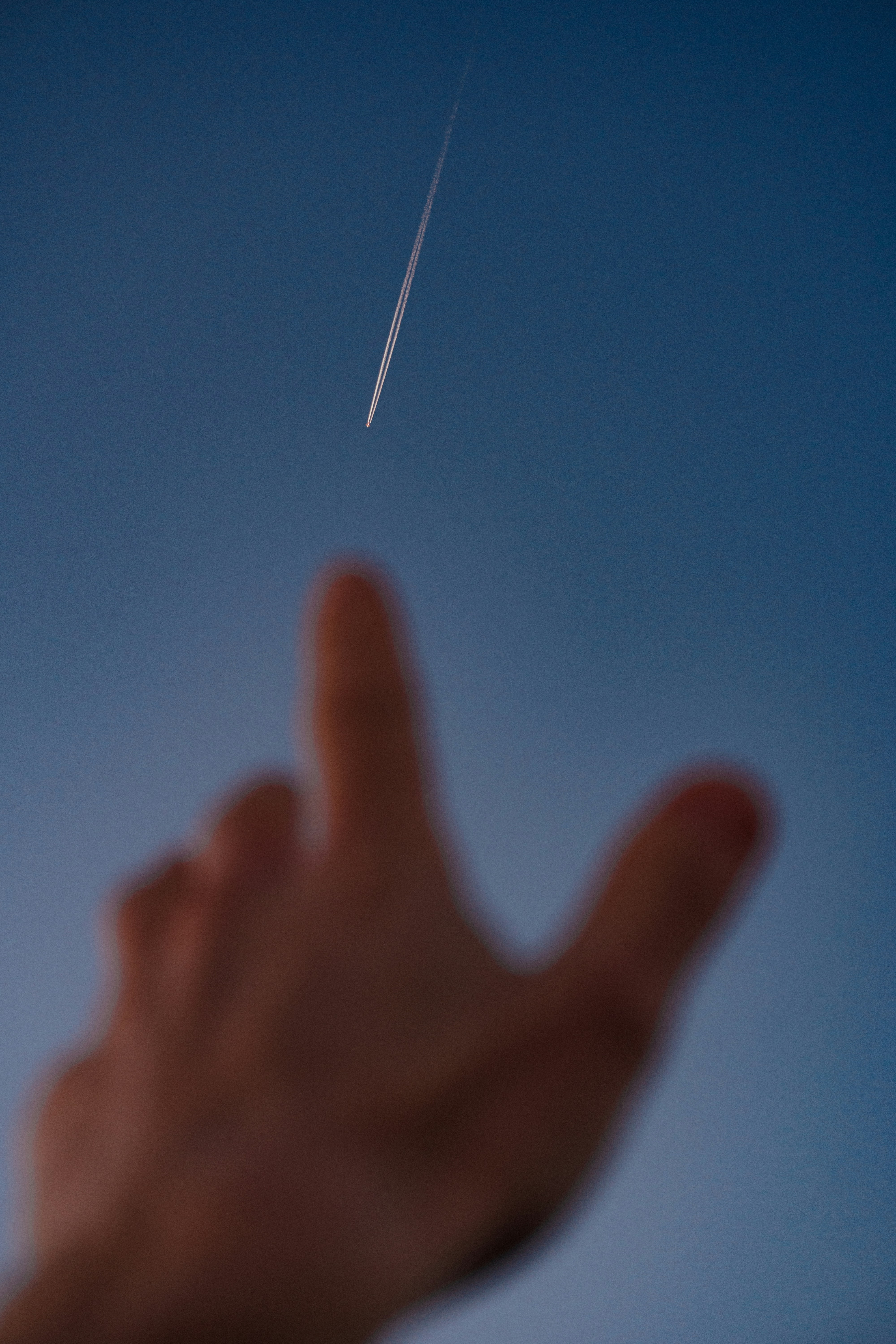 Hand reaching towards a distant airplane contrail
