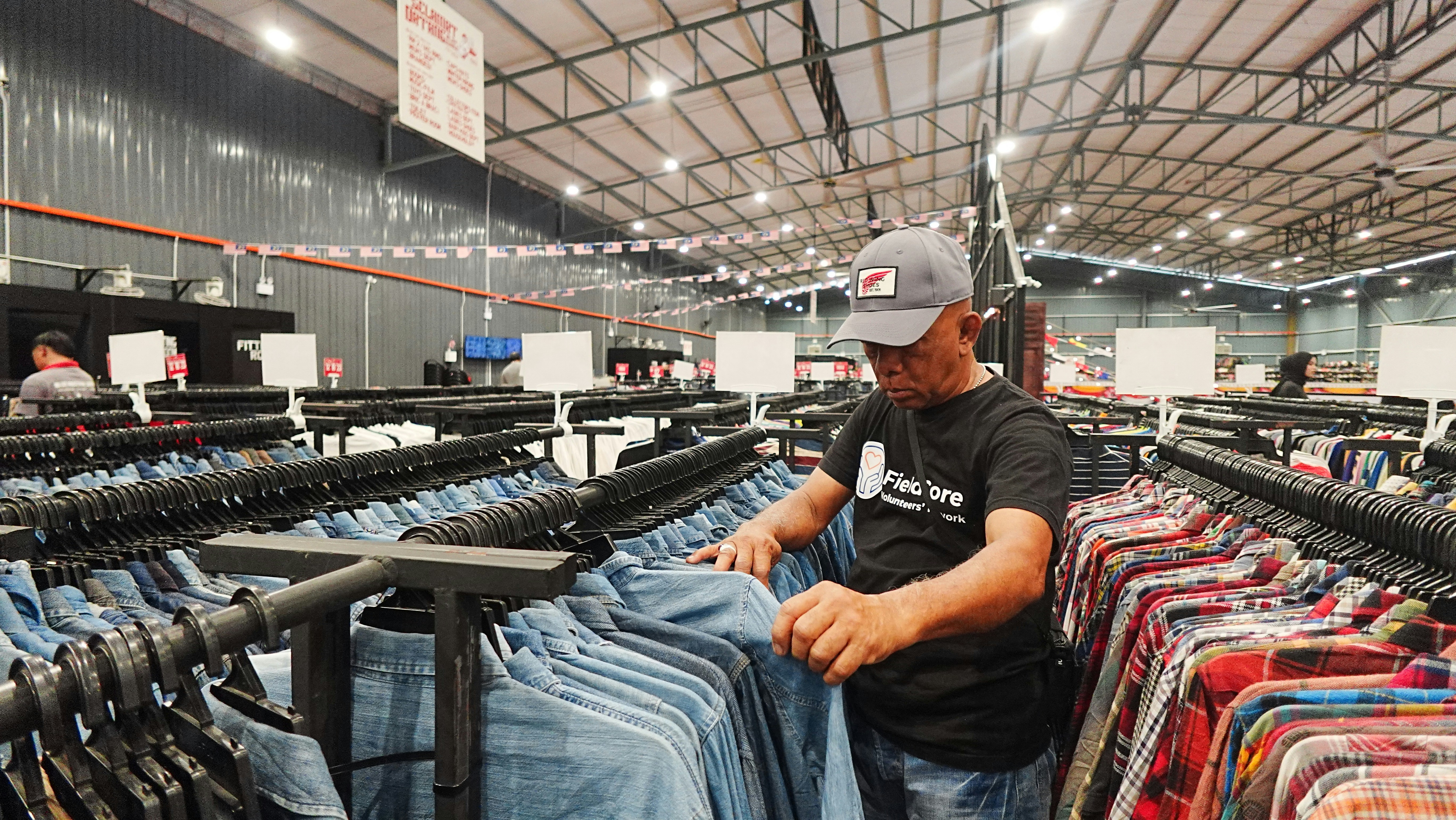 A man stands among clothing racks in a large warehouse, folding denim jeans on a rail. The scene emphasizes retail storage and manual handling in a bright, industrial setting.