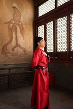 Man in red traditional chinese attire stands by mural.