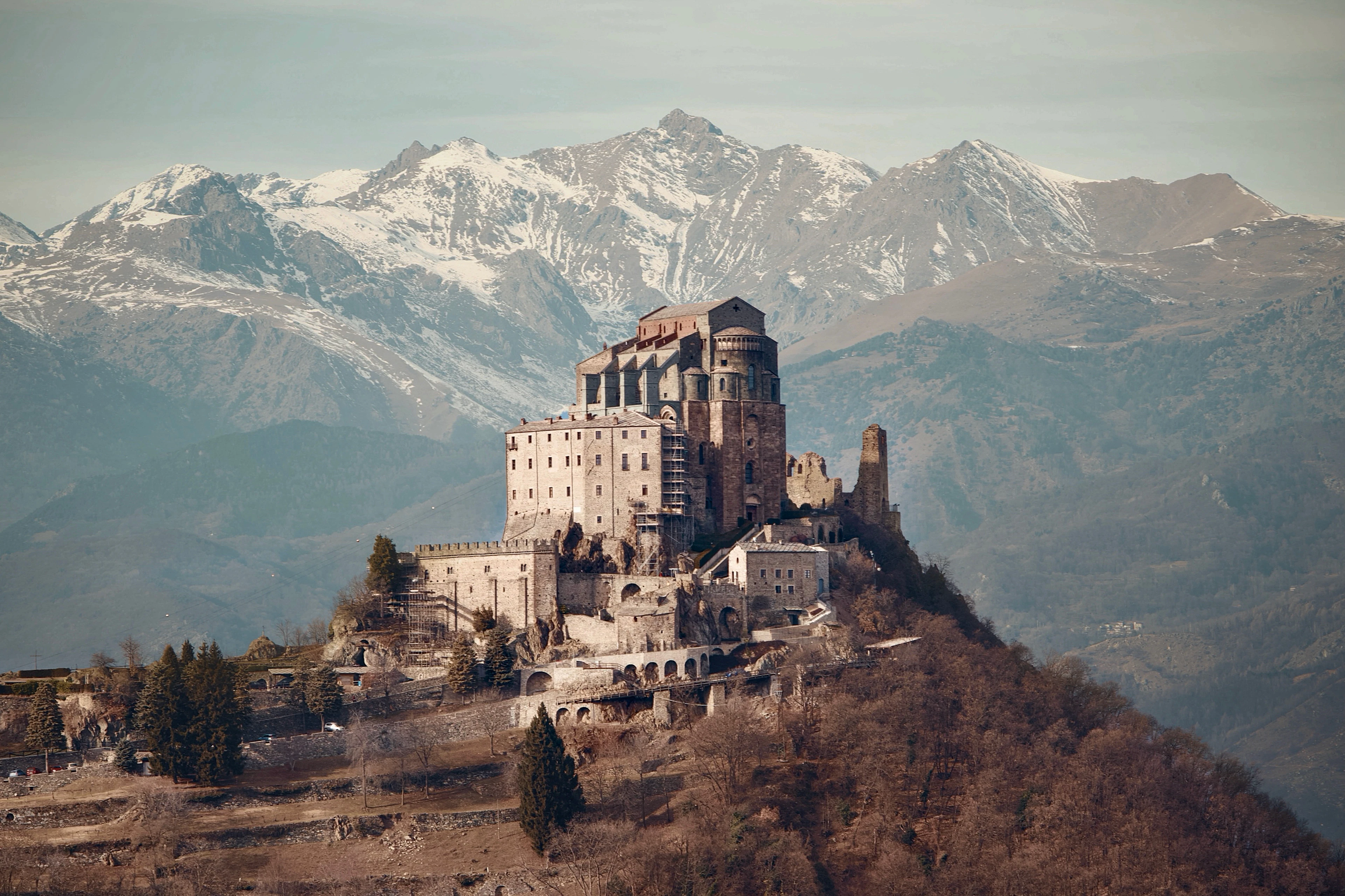 The Sacra di San Michele abbey rising from a rocky peak above the Susa Valley west of Turin, its pale stone walls and ruined tower standing against a wall of snow-covered Alps.